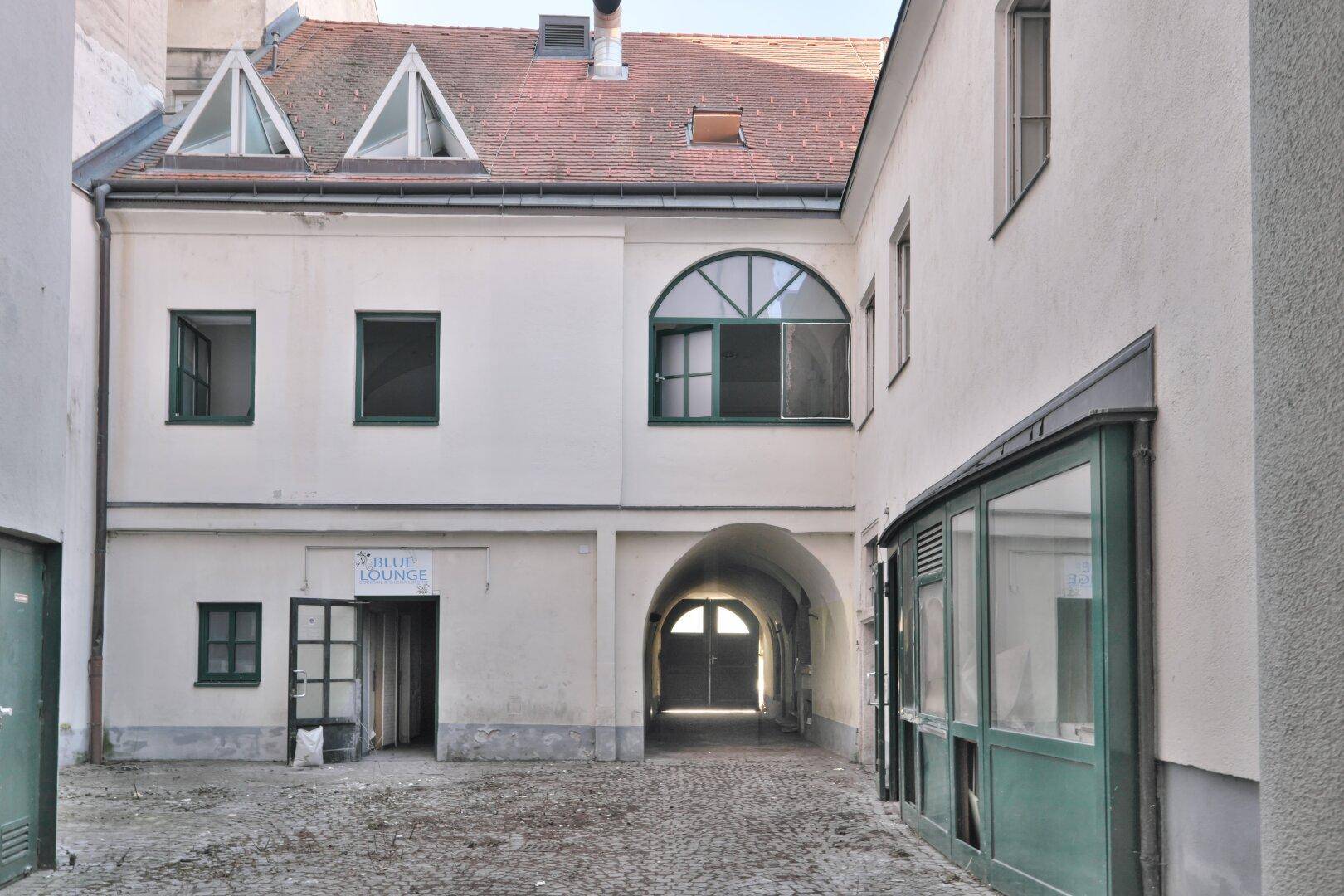 Innenhof mit Kopfsteinpflaster und einem Durchgang, umgeben von Gebäuden mit grünen Fensterrahmen.