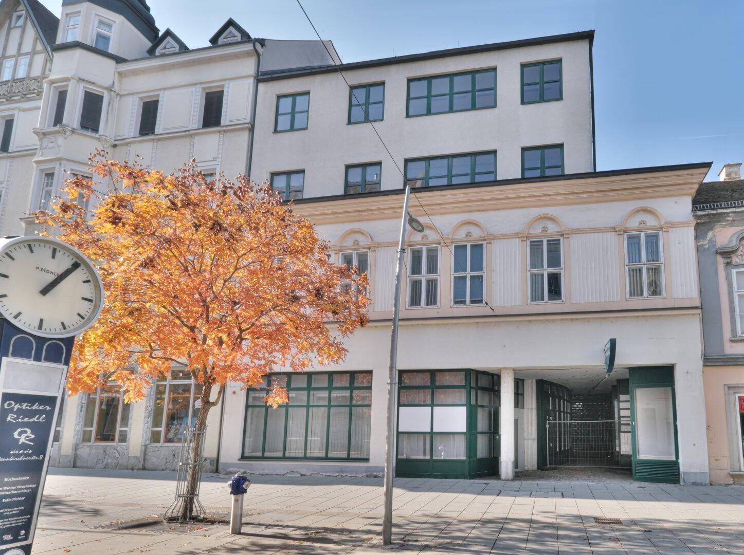 Historisches Gebäude mit Geschäftsfassade an einer belebten Straße, umgeben von Herbstbäumen und einem Uhrturm.