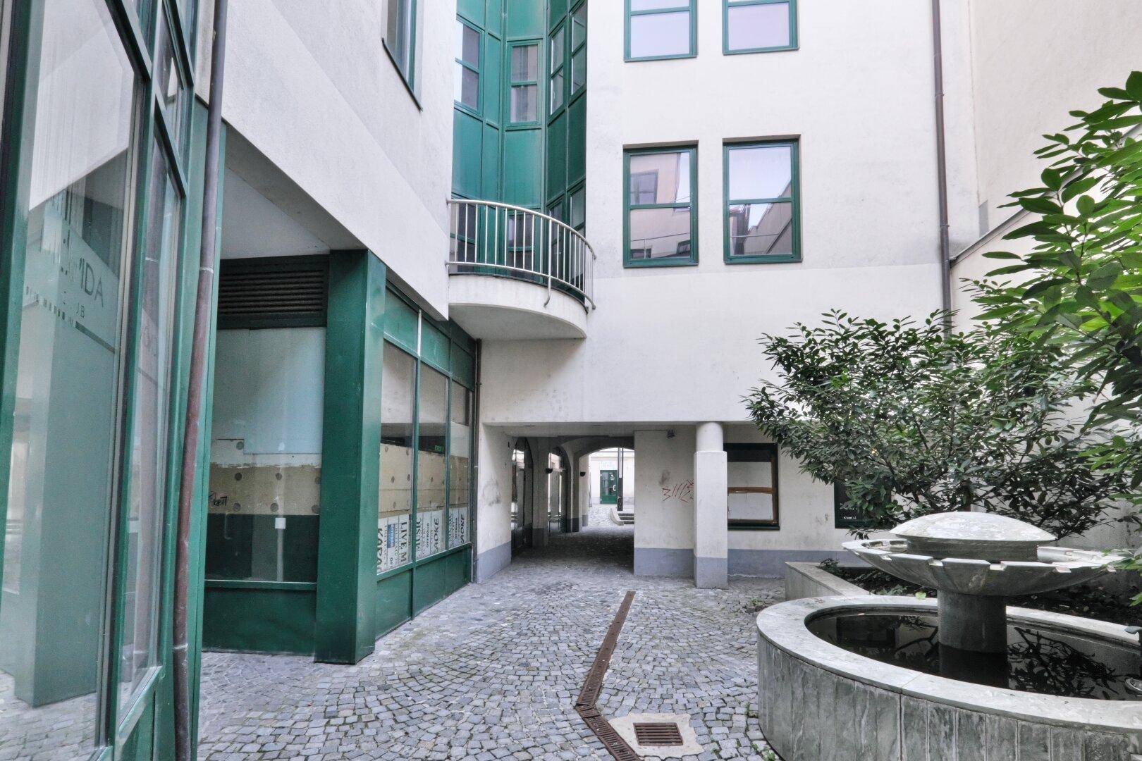 Innenhof mit einem Steinbrunnen und Kopfsteinpflaster, umgeben von Gebäuden mit grünen Fensterrahmen.