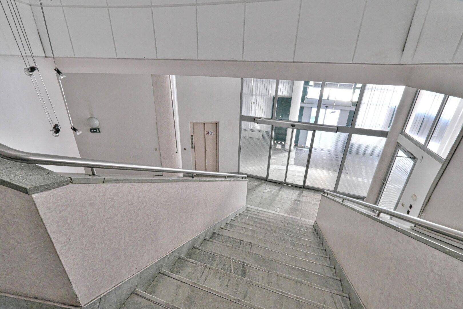 Weitläufige Marmortreppe mit Metallgeländer, die zu einem hellen Eingangsbereich mit Glasfront führt.
