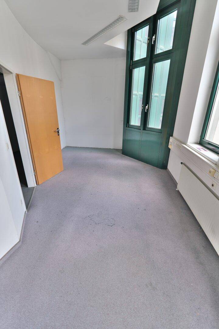 Kleiner Büroraum mit grauem Teppichboden, weißen Wänden und hohen grünen Fenstern.