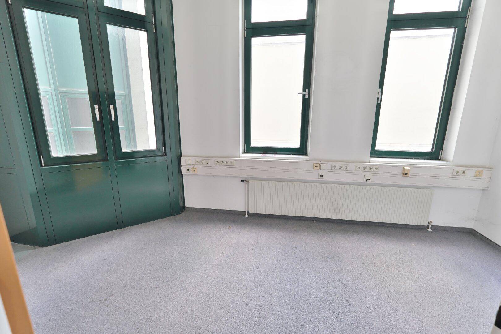 Kleiner Büroraum mit grauem Teppichboden, weißen Wänden und hohen grünen Fenstern mit Heizkörpern.