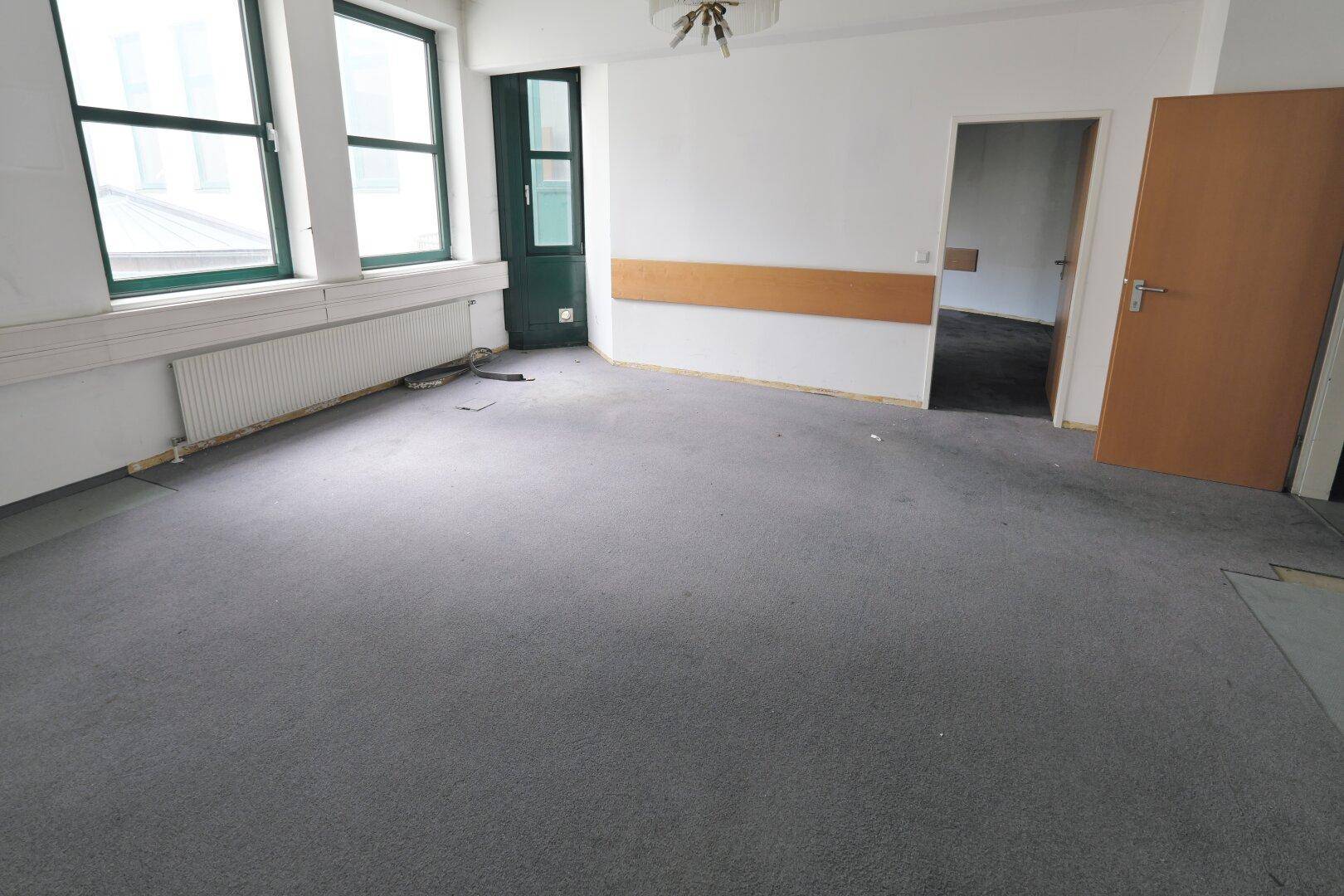 Leerer Büroraum mit grauem Teppichboden, weißen Wänden und großen Fenstern mit Heizkörpern darunter.