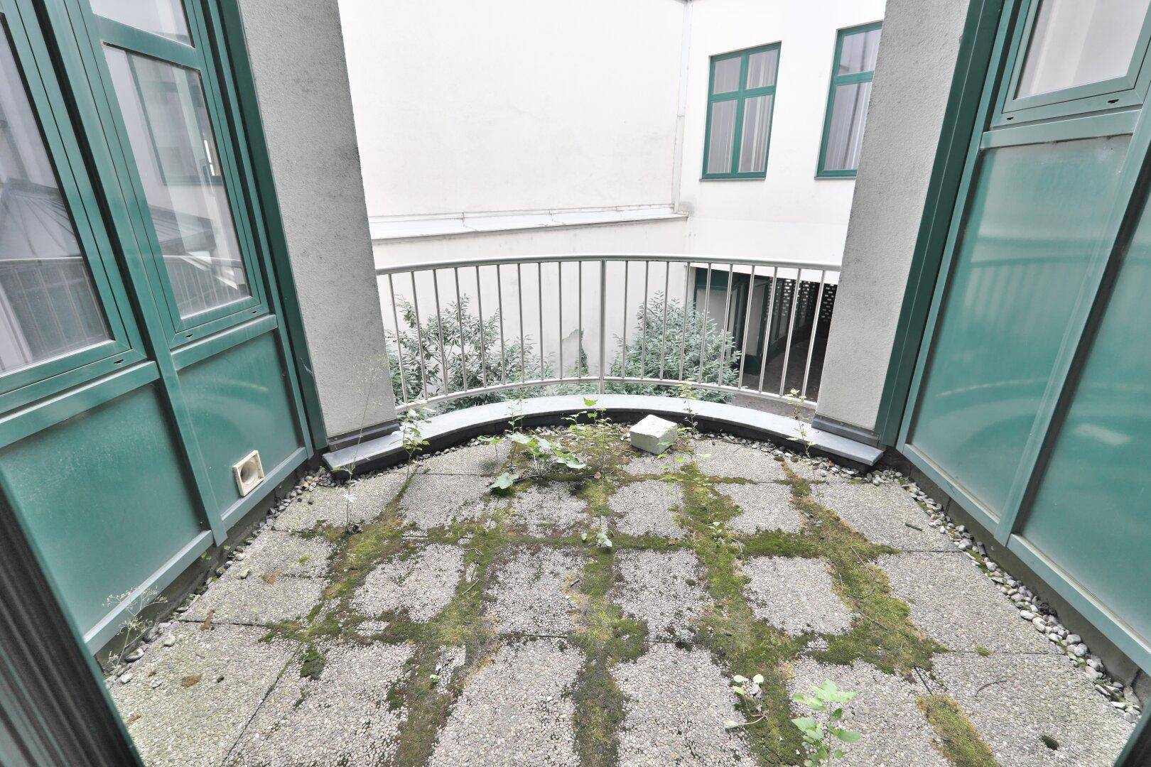 Kleiner Balkon mit Betonboden und Moosbewuchs, umgeben von grünen Fensterrahmen und einem Metallgeländer.