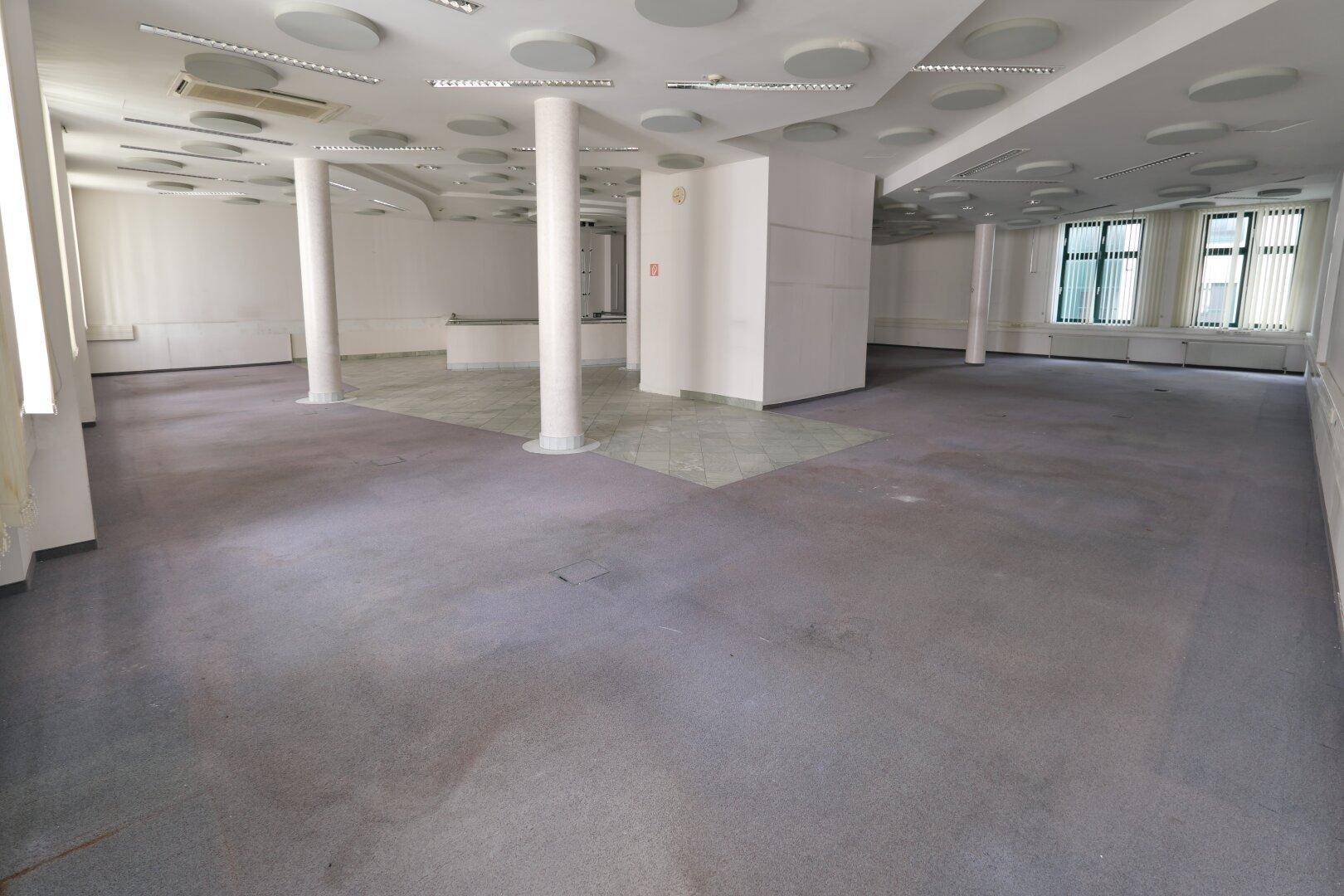 Geräumiger, leerer Büroraum mit grauem Teppichboden, vielen Säulen und einer Decke mit runden Leuchten.