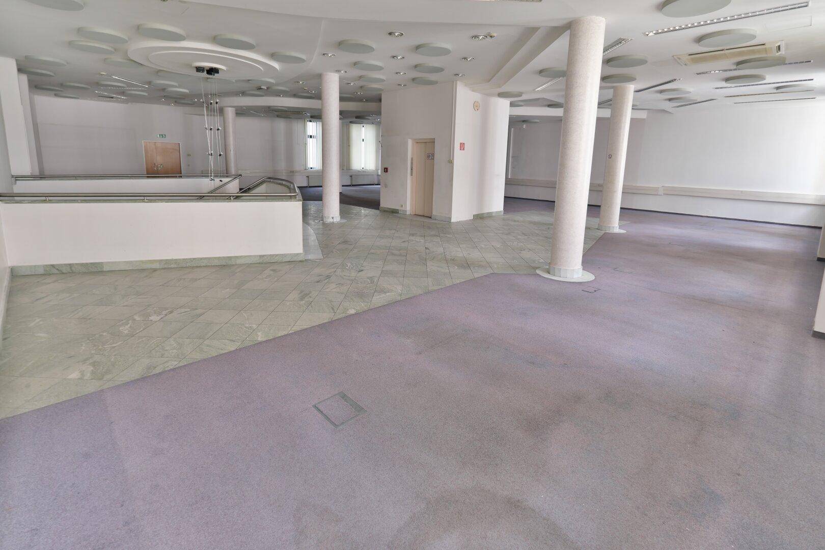 Großer, offener Büroraum mit Teppichboden, Säulen und einer Decke mit integrierten Leuchten.