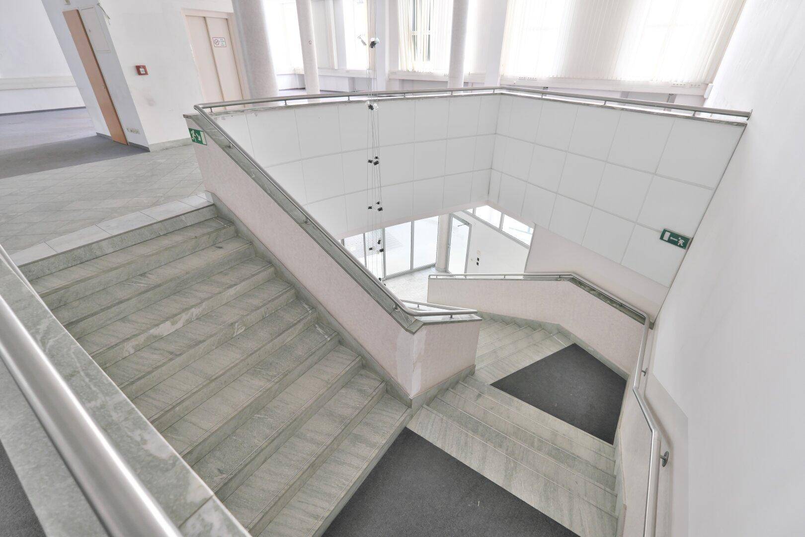 Weitläufige Marmortreppe mit Metallgeländer, die zu einem hellen Eingangsbereich mit Glasfront führt.