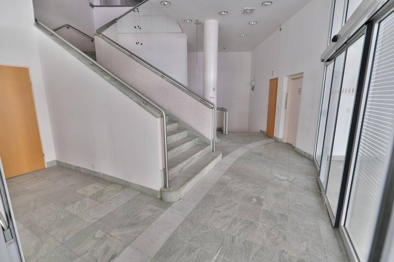Geräumiger Eingangsbereich mit Fliesenboden, einer Marmortreppe und automatischen Glastüren.