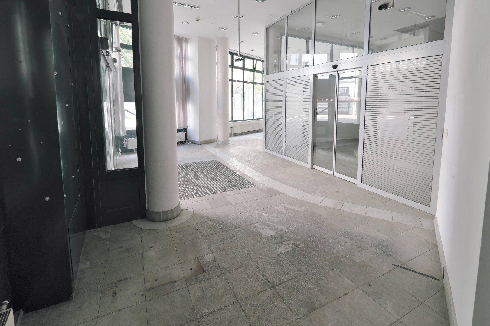 Heller Eingangsbereich mit Fliesenboden, automatischen Glastüren und Blick auf die Straße.
