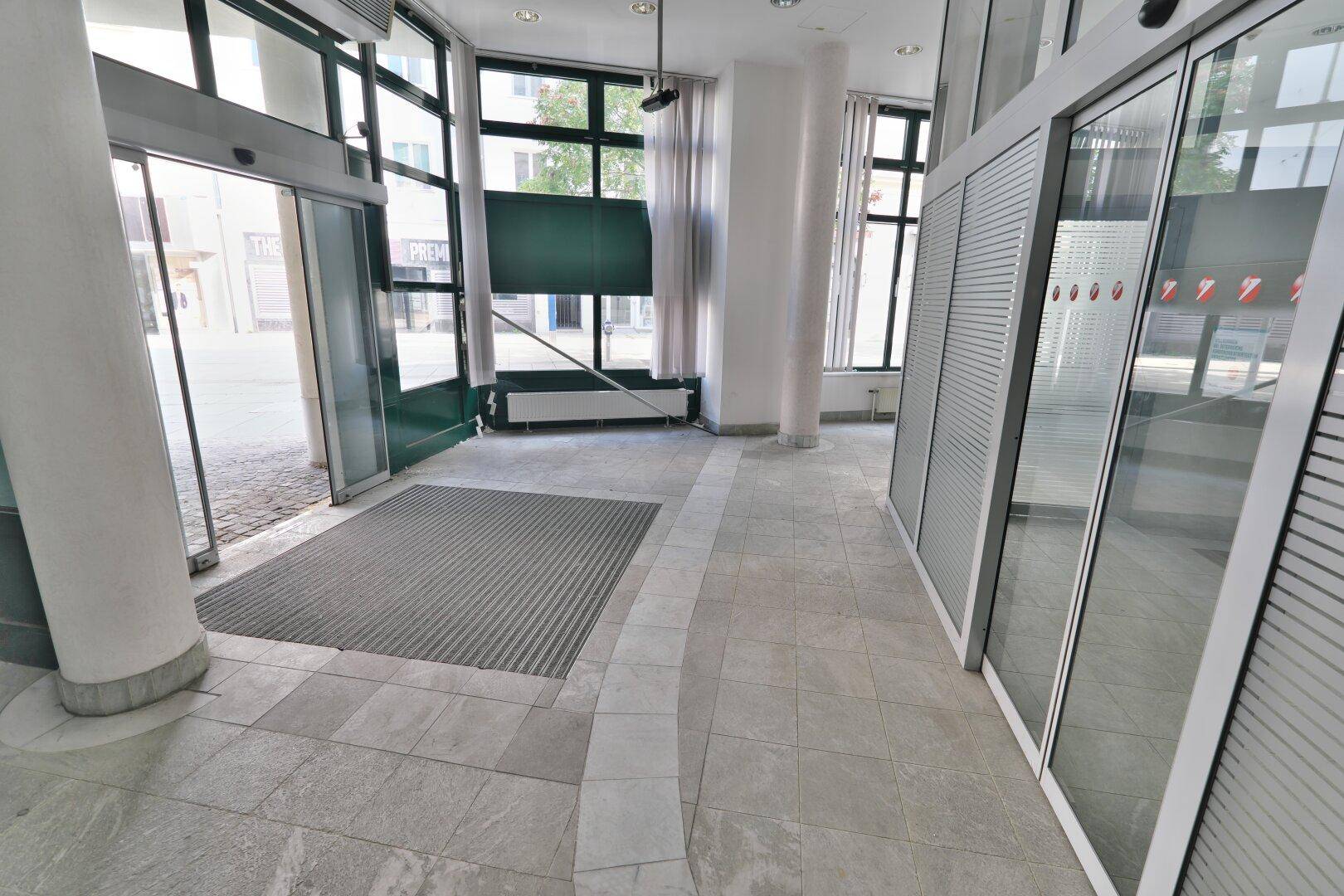 Heller Eingangsbereich mit Fliesenboden, automatischen Glastüren und Blick auf die Straße.