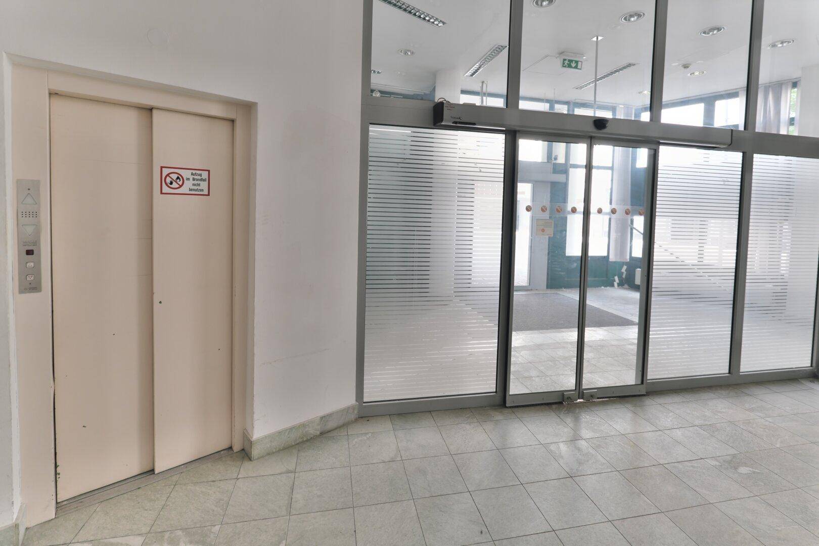 Eingangsbereich mit Aufzug und automatischen Glastüren, die zu einem hellen Flur mit Fliesenboden führen.