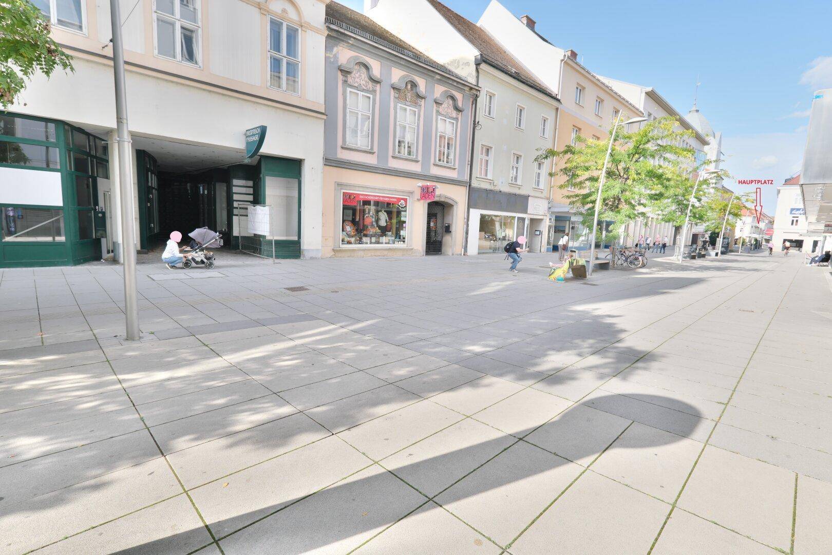 Belebtes Stadtbild mit Geschäften und Passanten auf einem breiten Bürgersteig an einem sonnigen Tag.