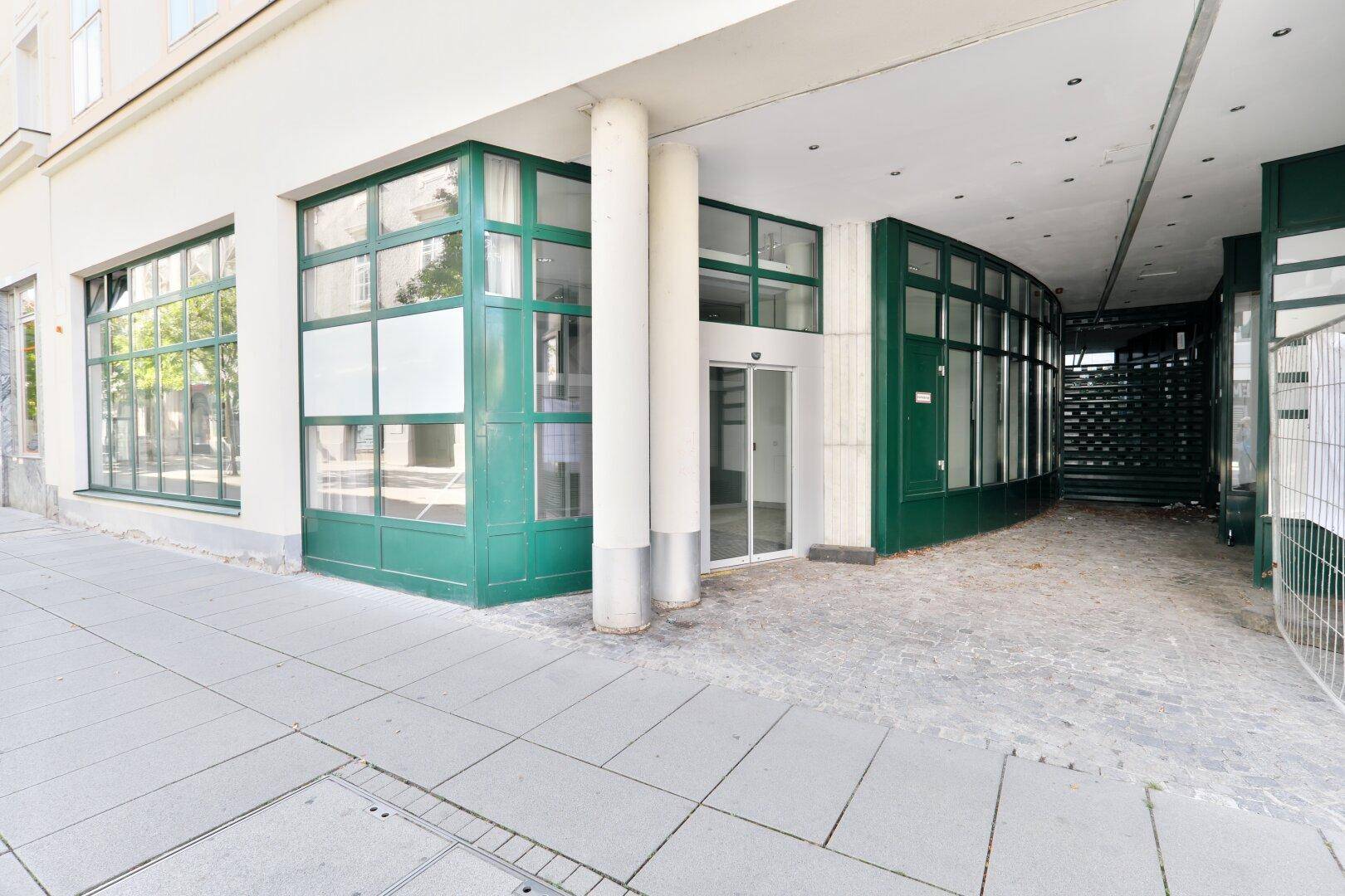 Eingangsbereich eines Geschäftsgebäudes mit großen Schaufenstern und grünen Fensterrahmen an einer belebten Straße.