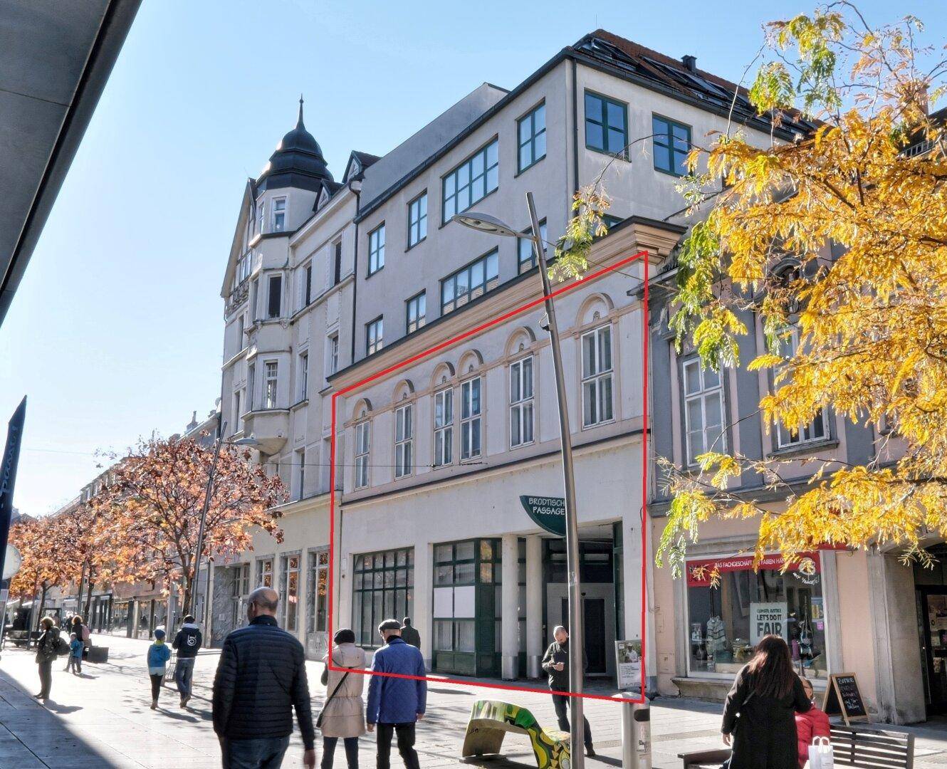 Historisches Gebäude mit markierter Fassade an einer belebten Einkaufsstraße mit Herbstbäumen.