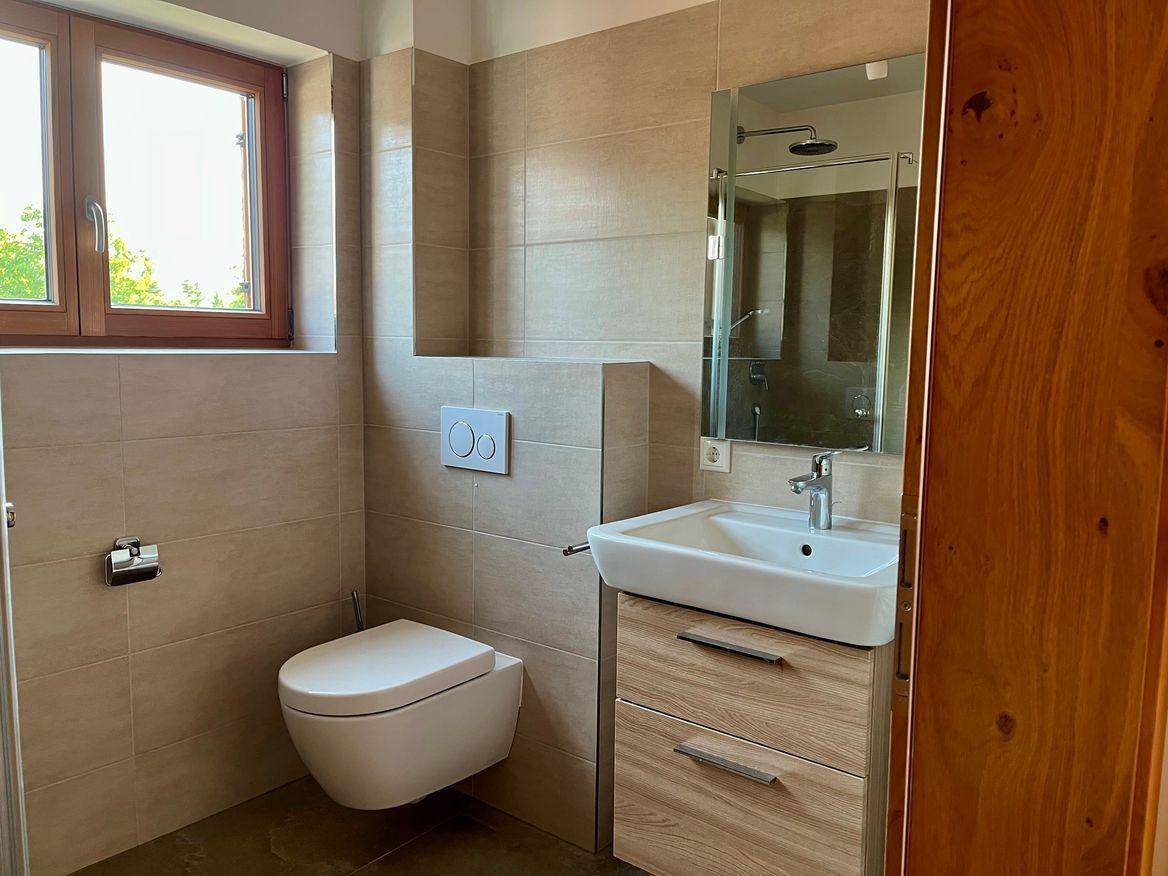 Modernes Badezimmer mit Wand-WC, Waschtisch, Dusche und Fenster für natürliche Belüftung.