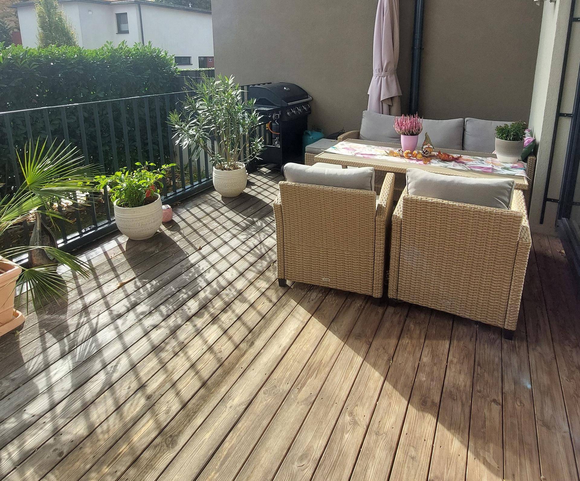 Sonnige Holzterrasse mit bequemen Gartenmöbeln, Pflanzen und Grillbereich, ideal zum Entspannen.