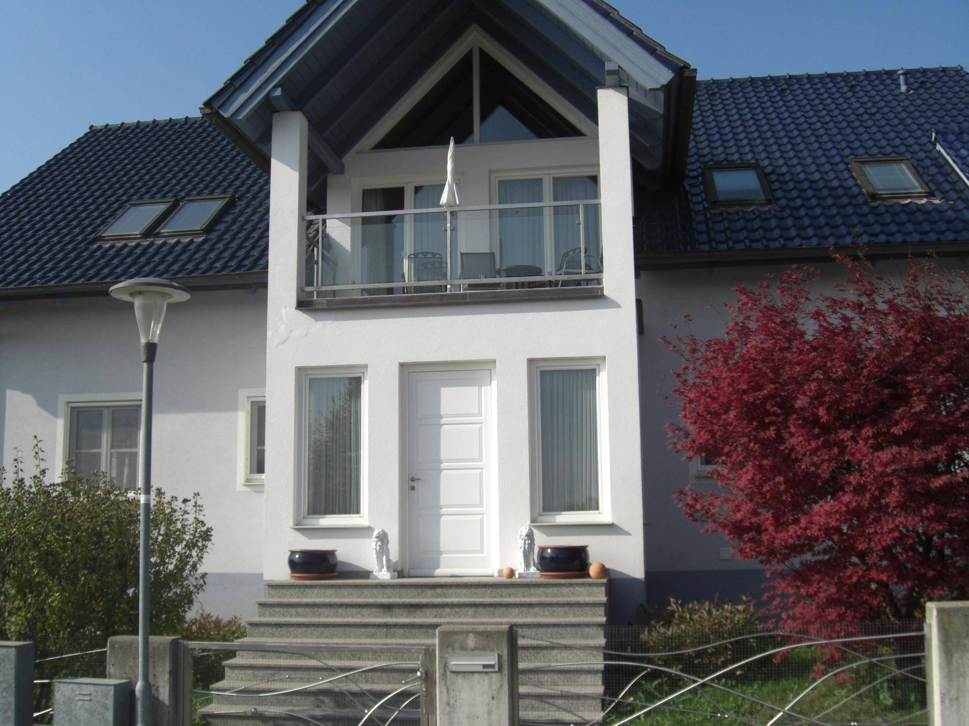 Vorderansicht des Hauses mit weißer Fassade, Eingangsbereich, Balkon und dunklem Ziegeldach unter blauem Himmel.