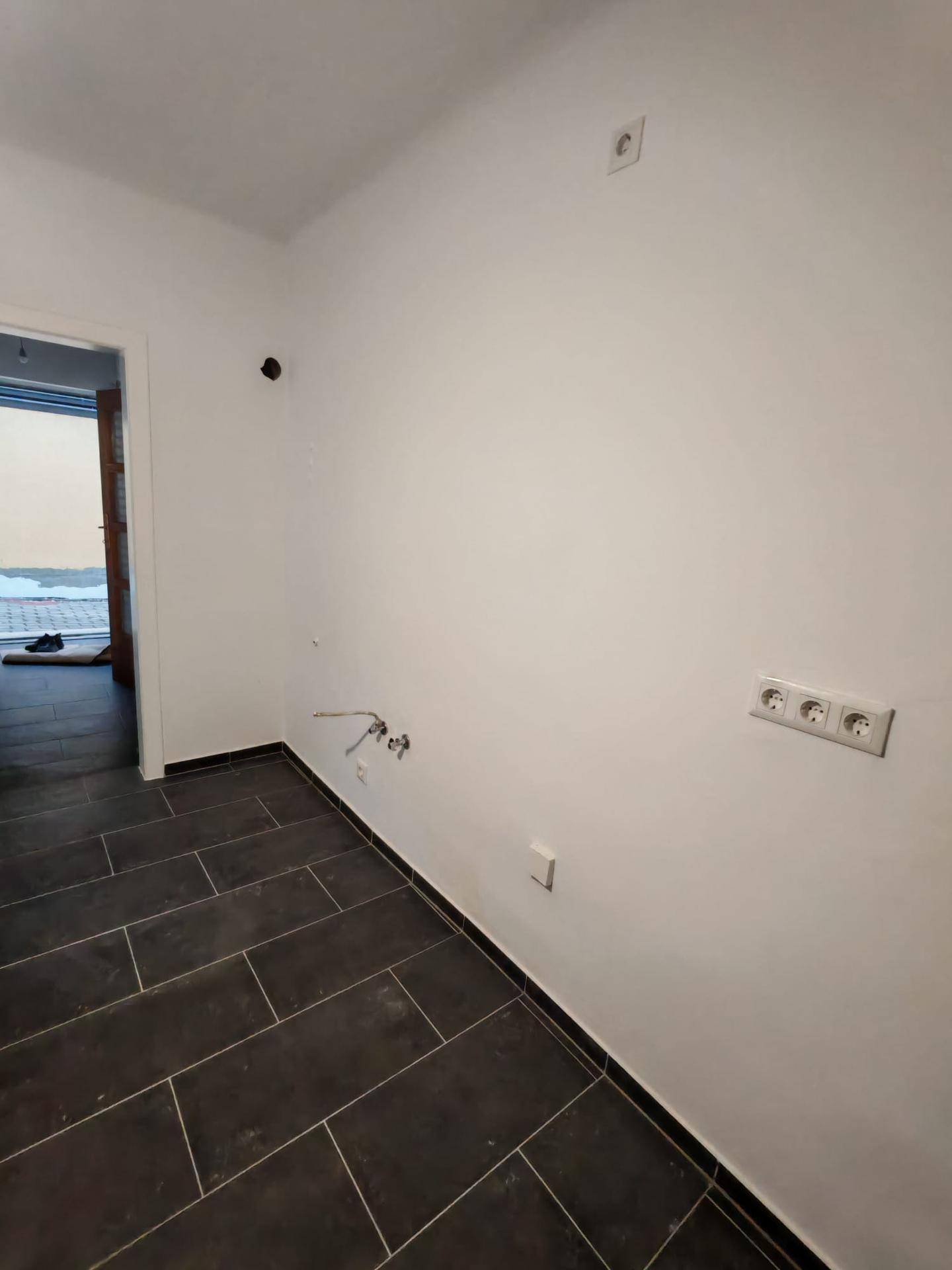 Geräumiger Küchenbereich mit dunklem Fliesenboden und vorbereiteten Installationen für Geräte.