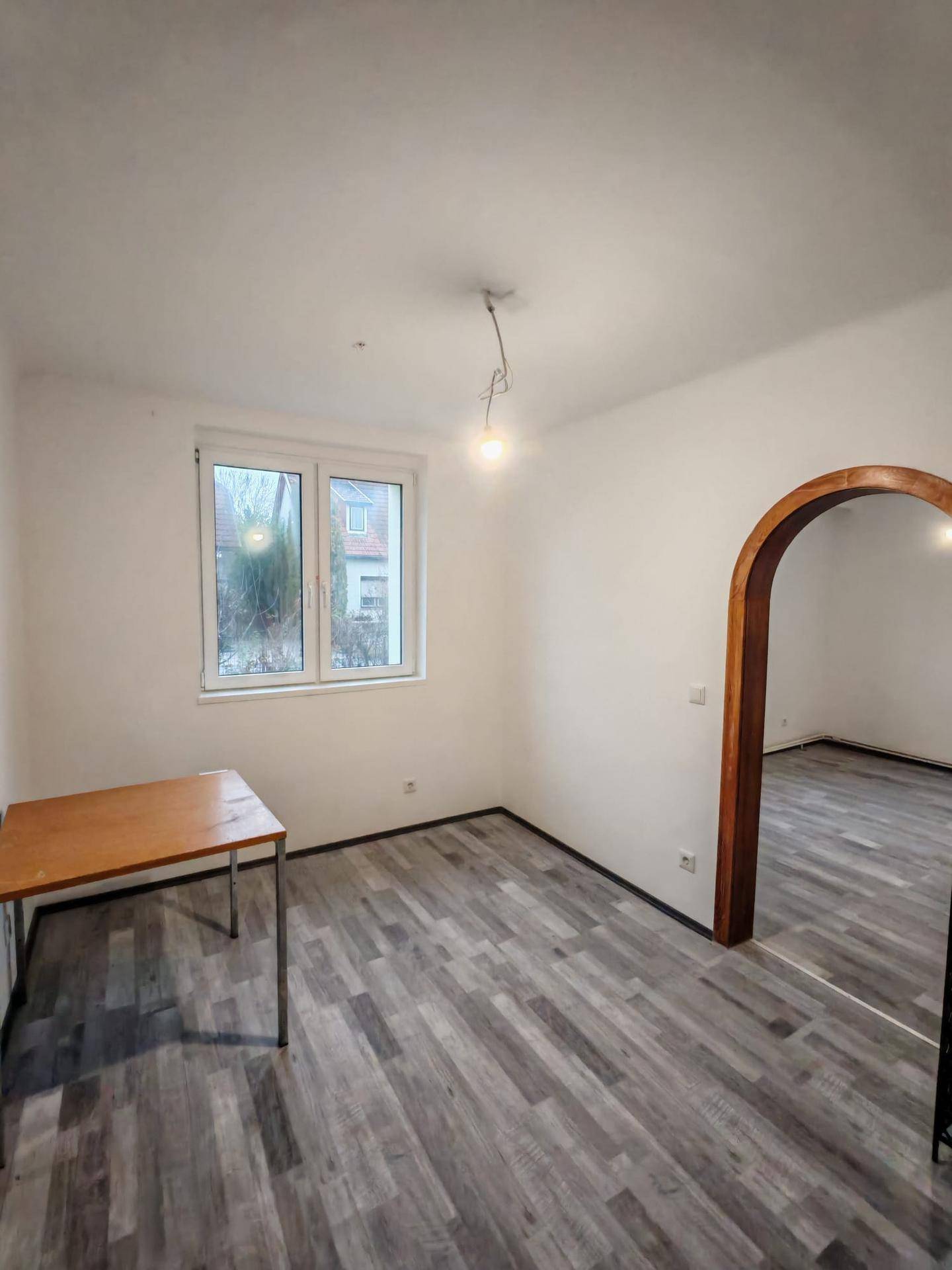 Leeres Zimmer mit hellem Laminatboden, einem Fenster und einem einfachen Holztisch.