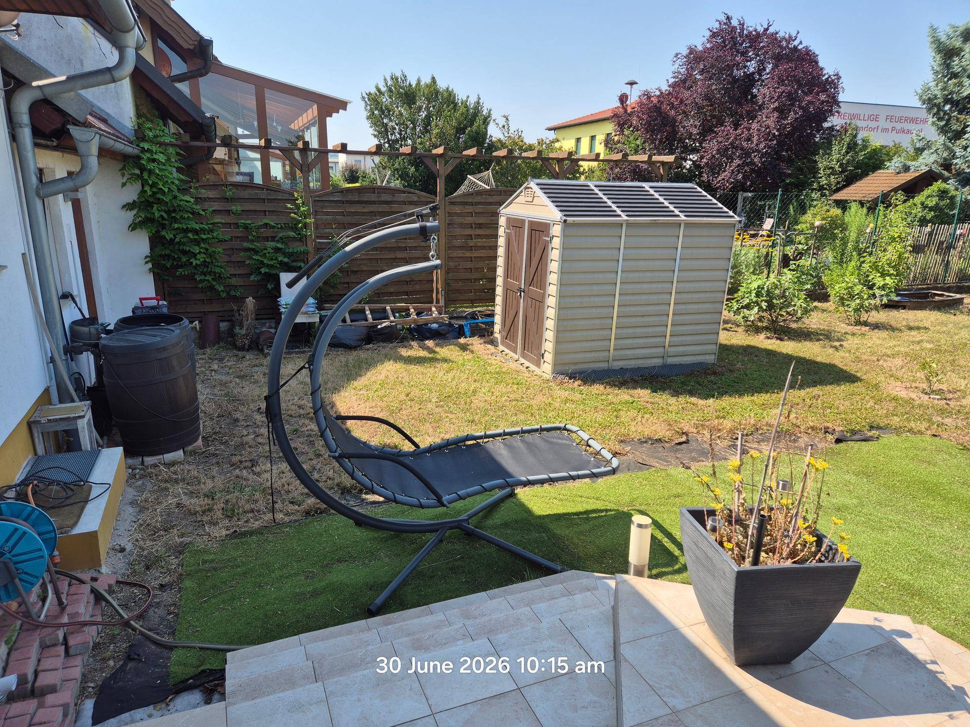Gepflegter Gartenbereich mit Gartenhaus, Schaukelstuhl und Rasenfläche an sonnigem Tag.