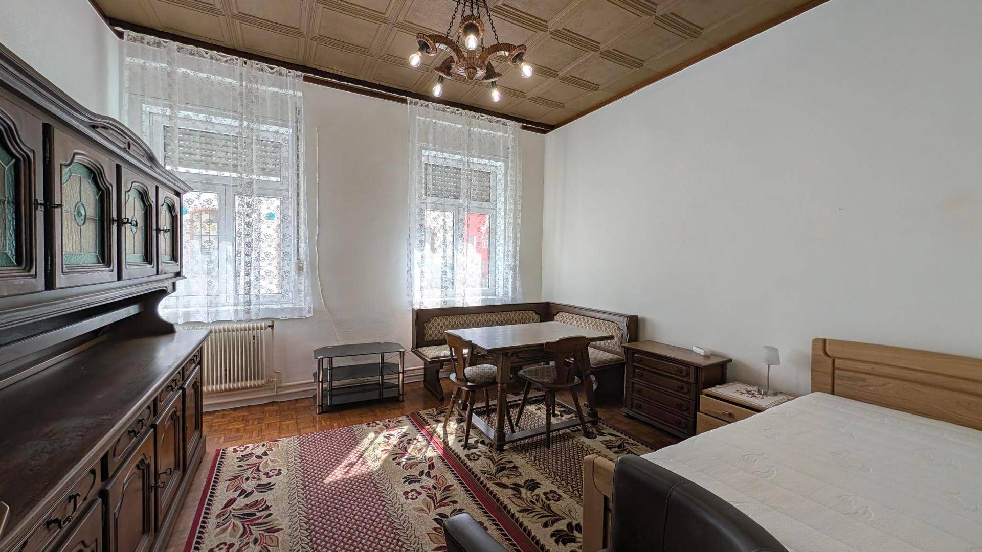 Geräumiges Schlafzimmer mit traditionellen Holzmöbeln, Essbereich und gemusterter Decke, gut beleuchtet durch große Fenster.