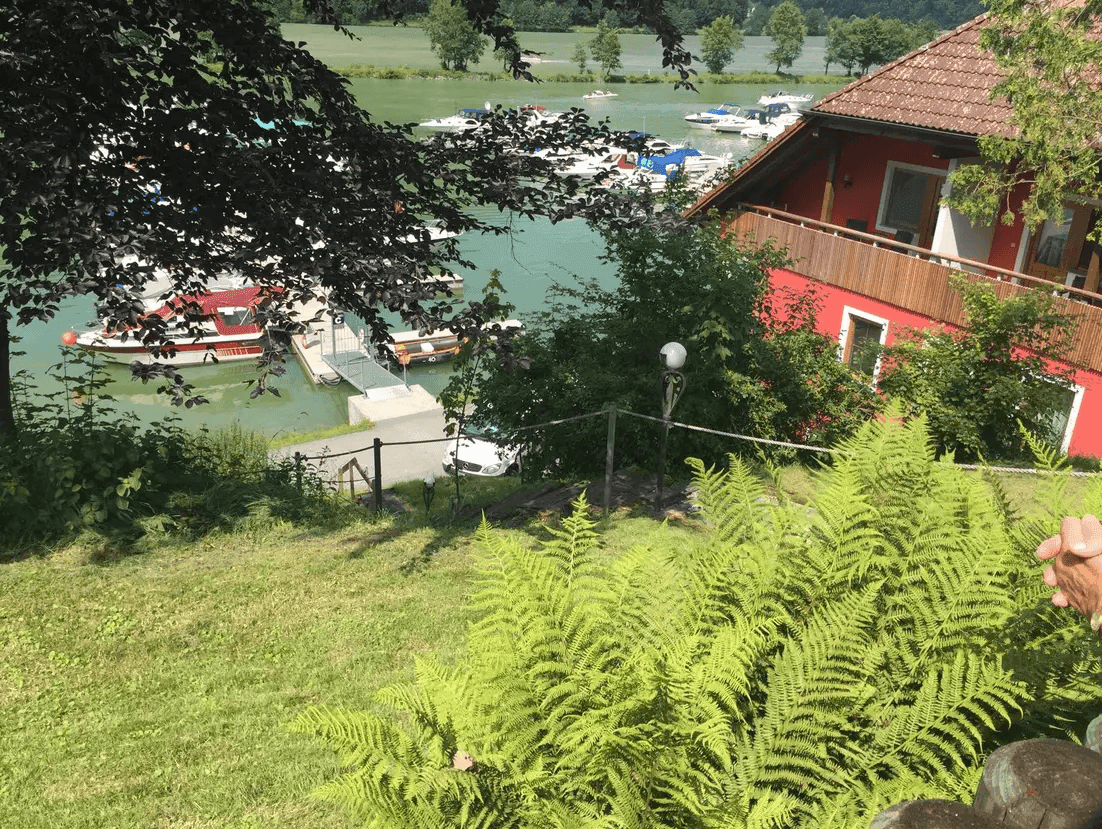 Blick auf das rote Haus am Seeufer mit Bootsanlegestellen und üppiger Vegetation.