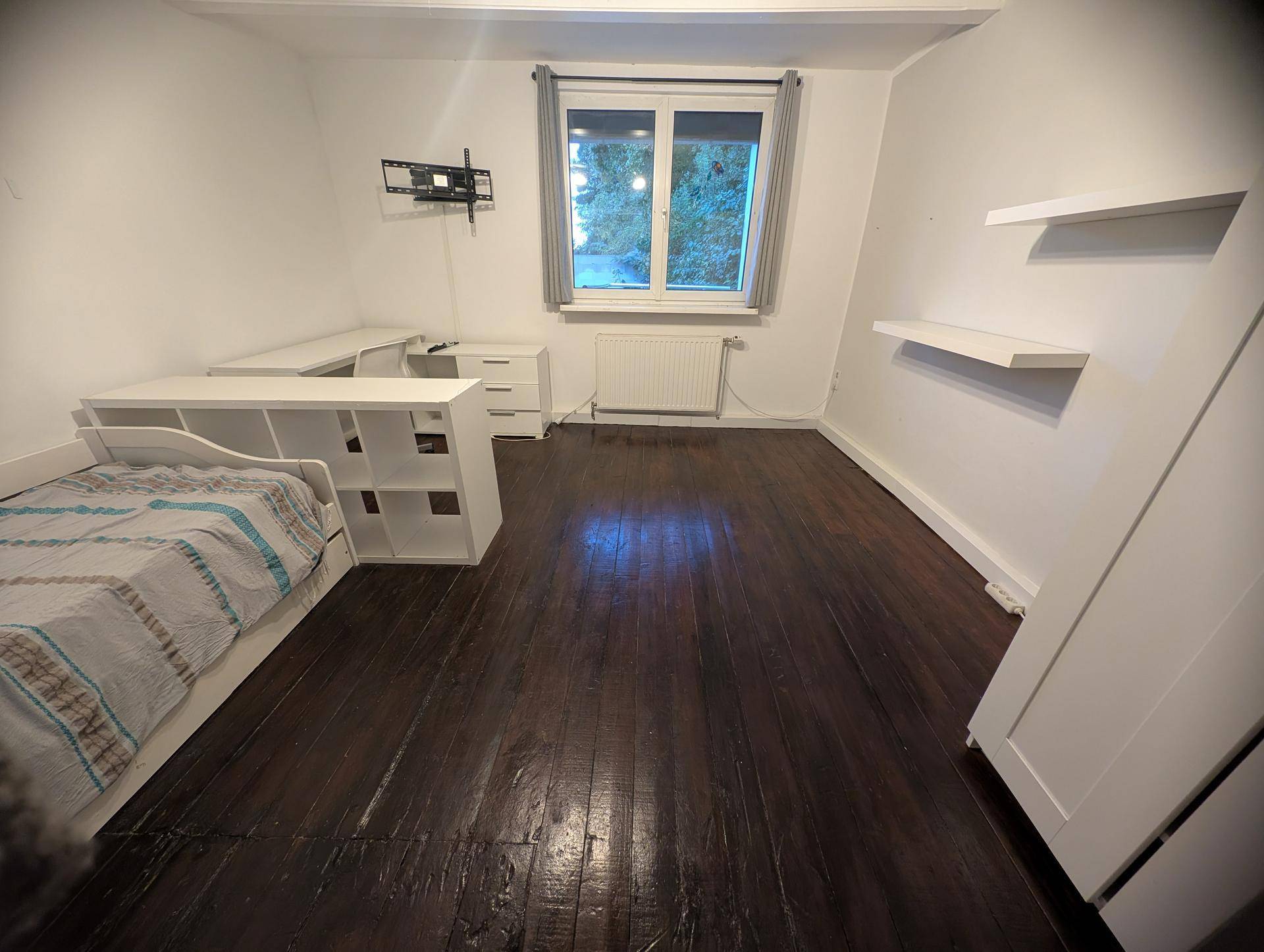 Helles Schlafzimmer mit dunklem Holzboden, Schreibtisch und Fenster mit Blick ins Grüne.
