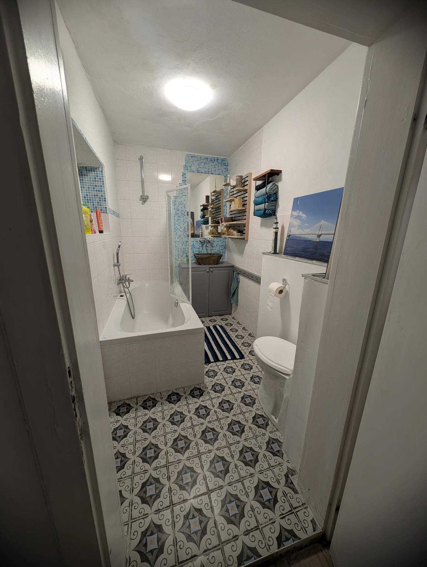 Kleines Badezimmer mit Badewanne, Duschfunktion und gemusterten Bodenfliesen in Blau-Weiß.