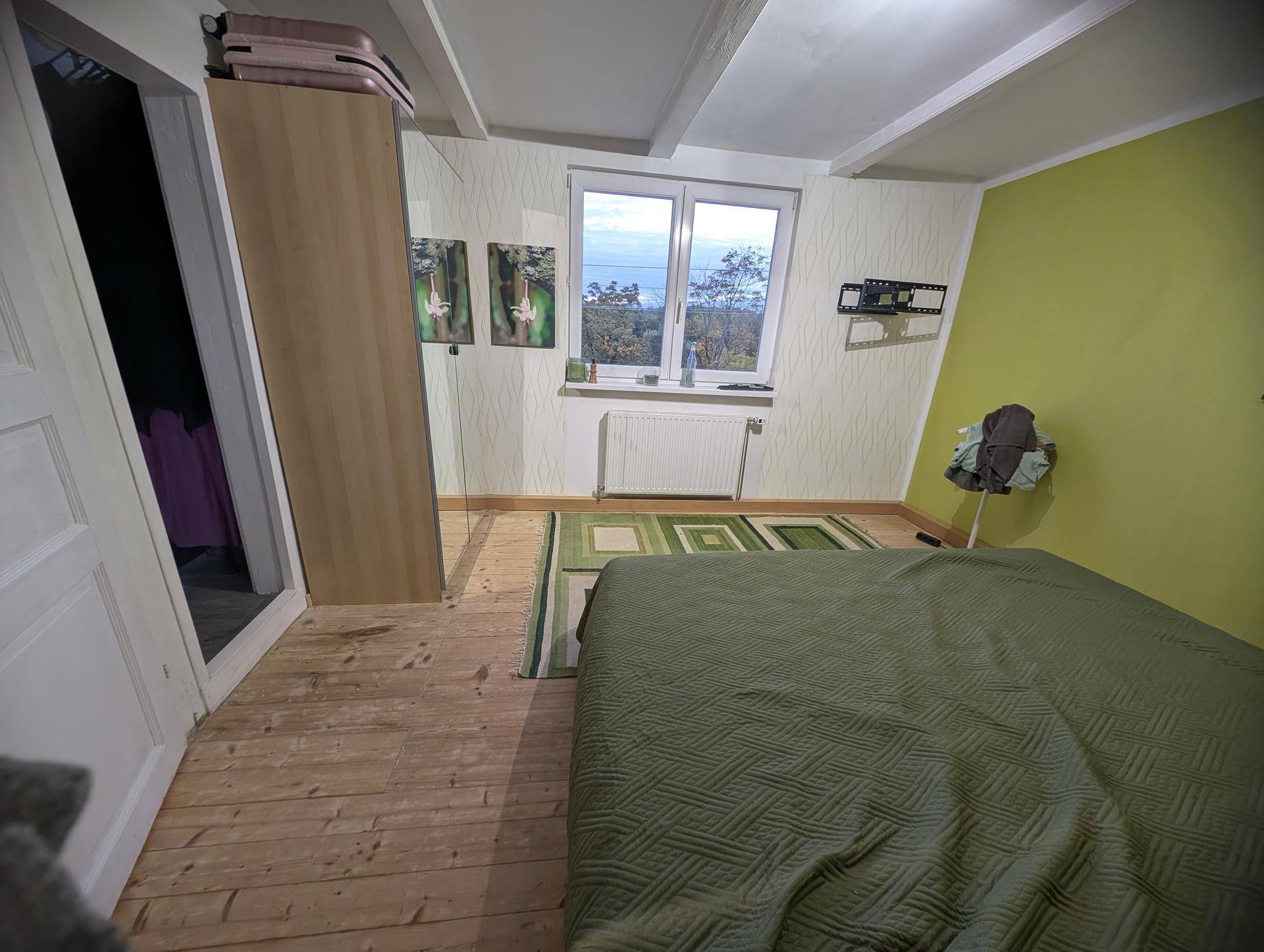 Schlafzimmer mit Holzboden, grüner Akzentwand und Fenster mit Blick auf die Natur.