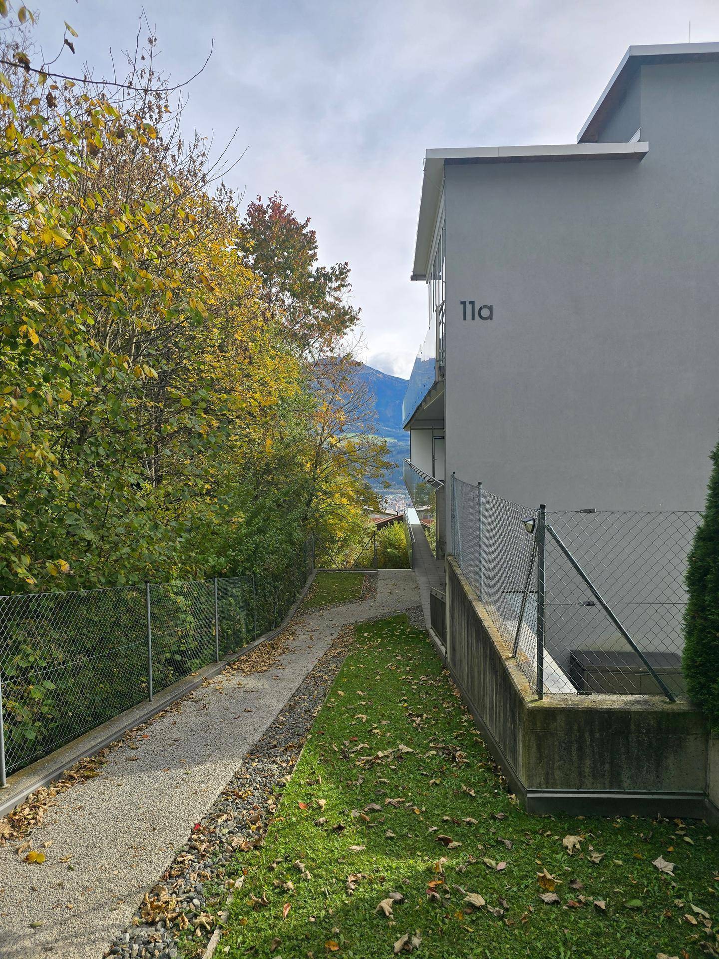 Seitenansicht des Gebäudes mit einem schmalen Weg und herbstlicher Vegetation entlang des Zauns.