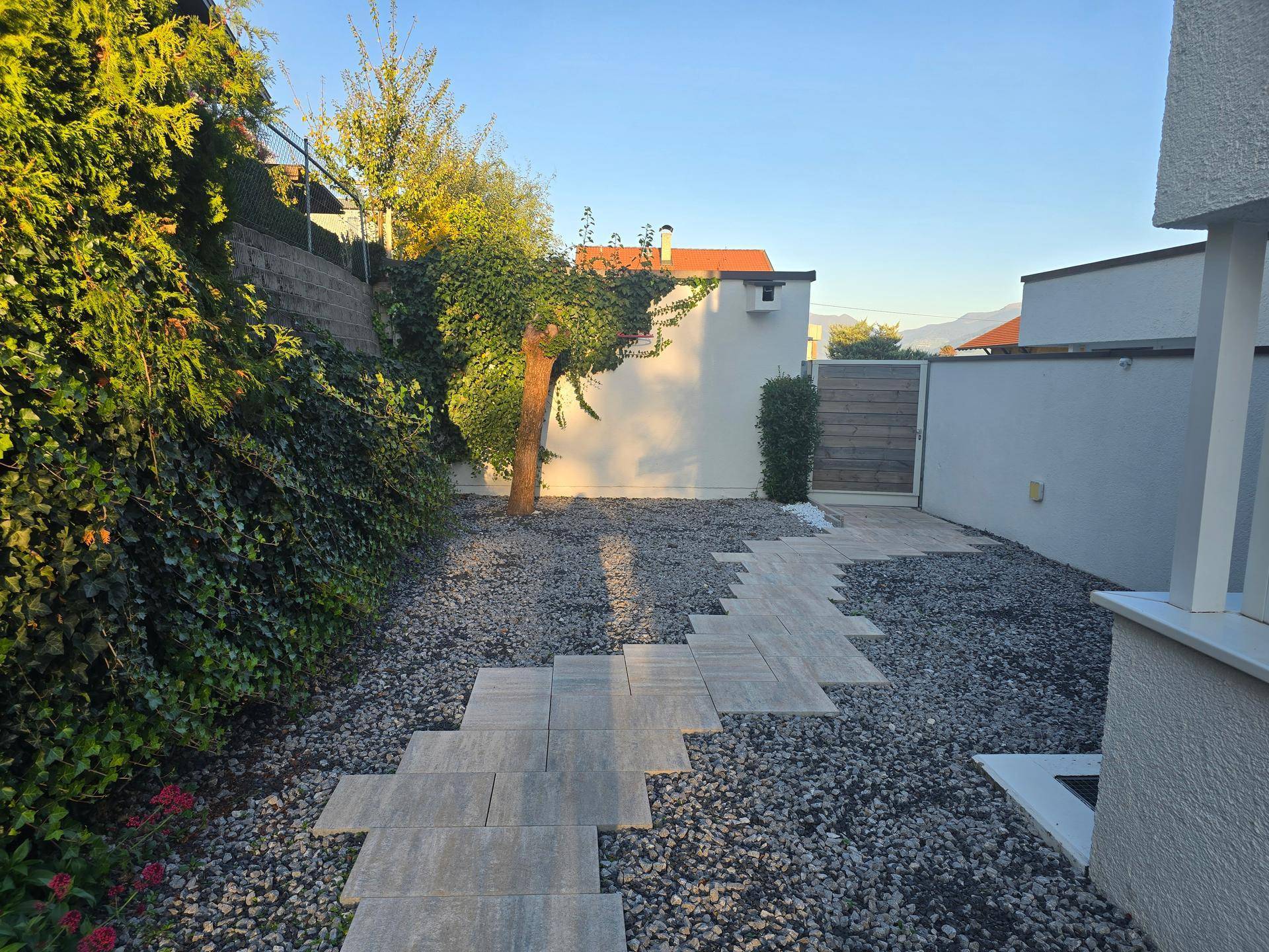 Gartenweg aus Steinplatten durch Kies, umgeben von üppigem Grün und einer hohen Hecke.