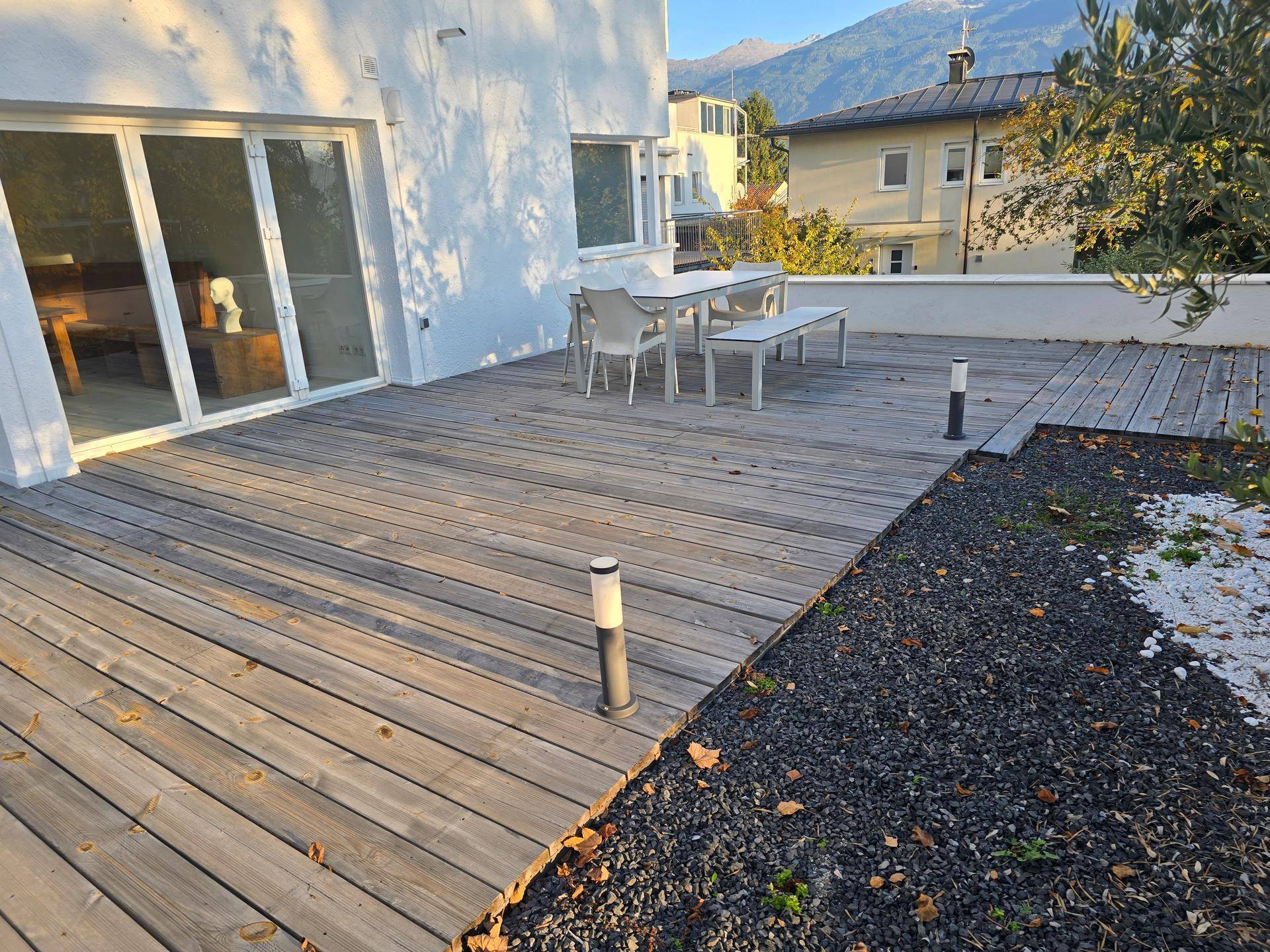 Geräumige Holzterrasse mit Esstisch und Sitzgelegenheiten, umgeben von Kies und Bergblick.