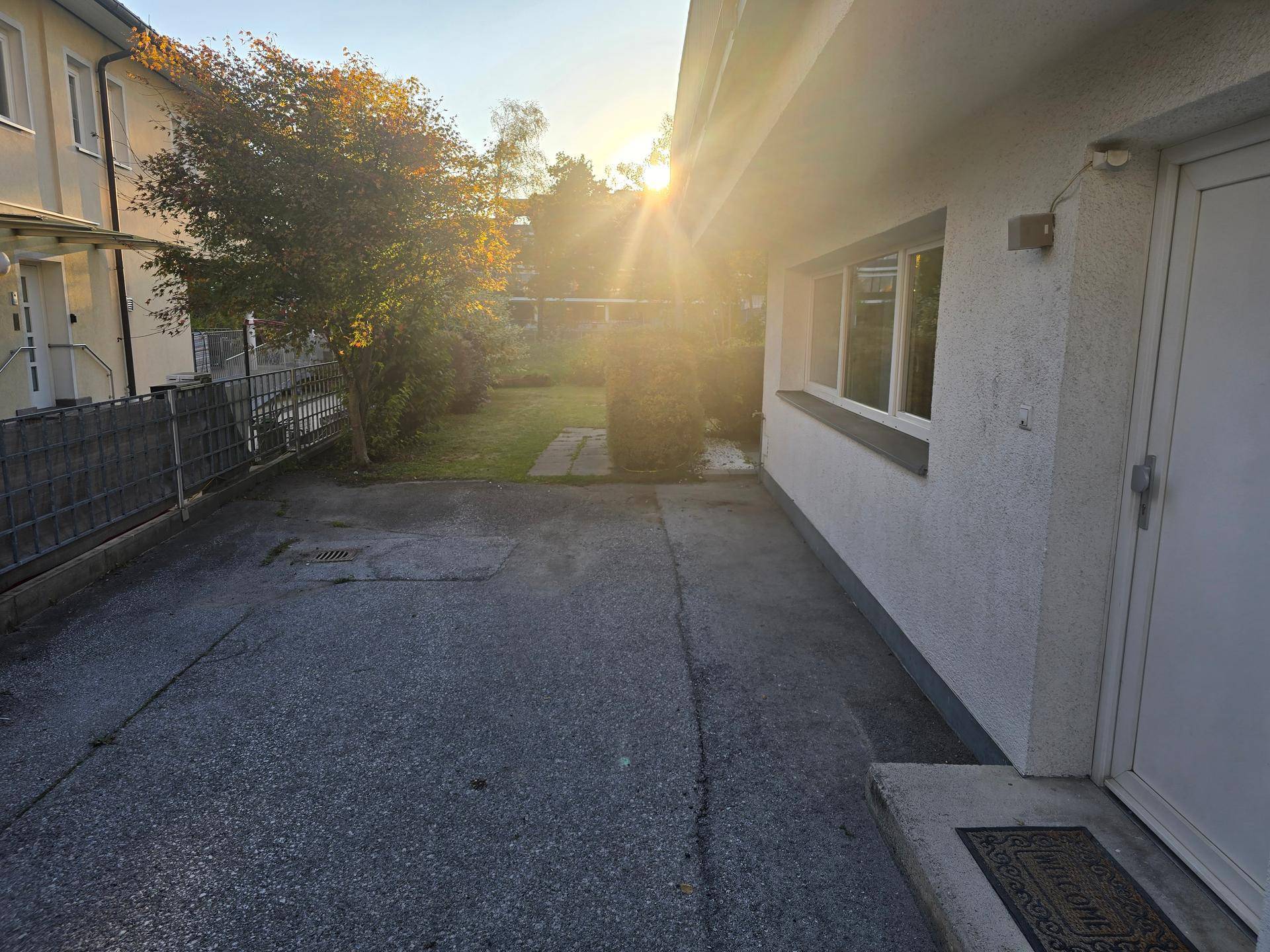 Einfahrt oder Parkplatz neben dem Haus, mit Zaun und Gartenbereich im Hintergrund.