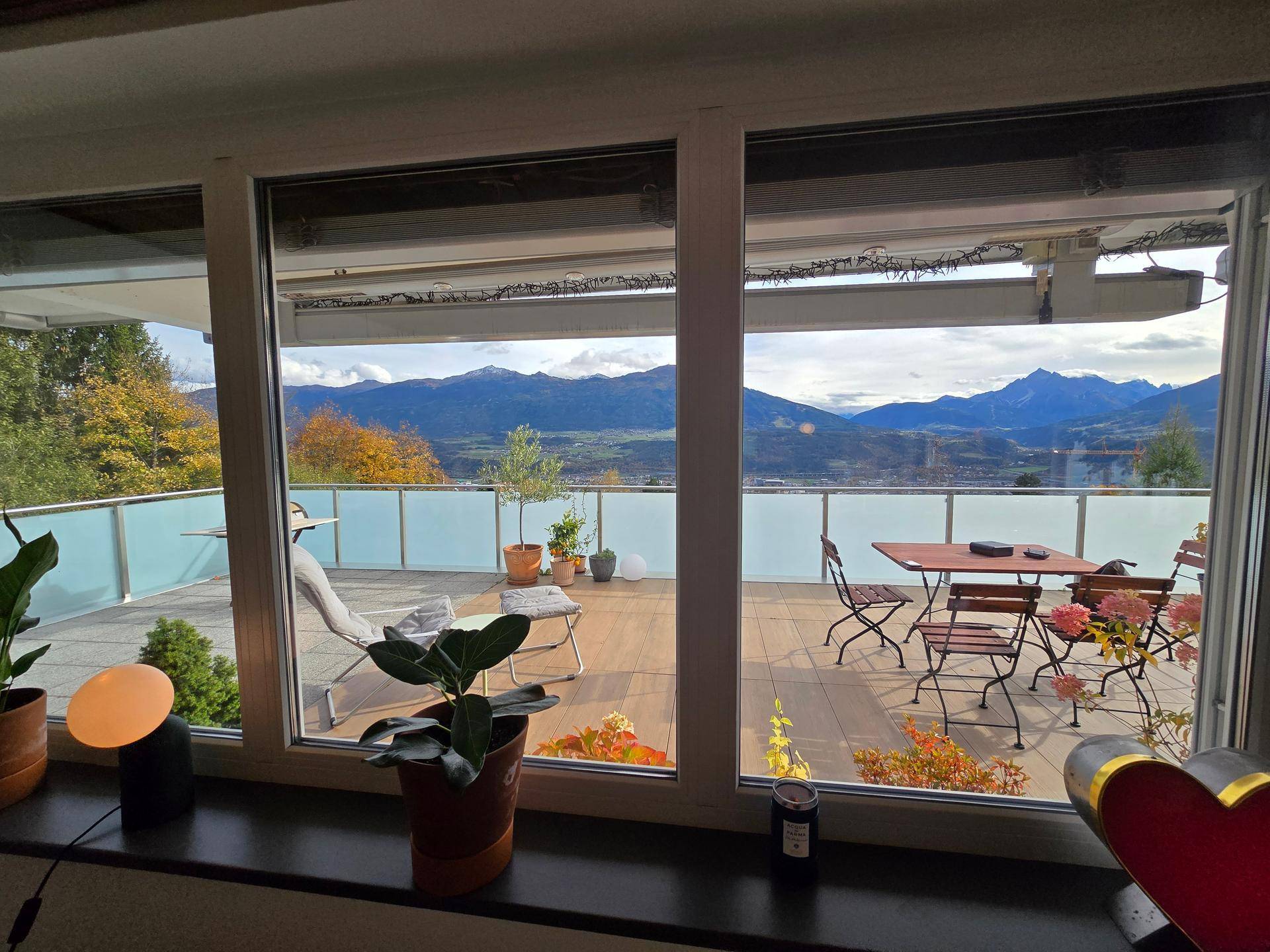 Innenansicht des Wohnbereichs mit großem Fenster und Blick auf die möblierte Terrasse und die Berge.