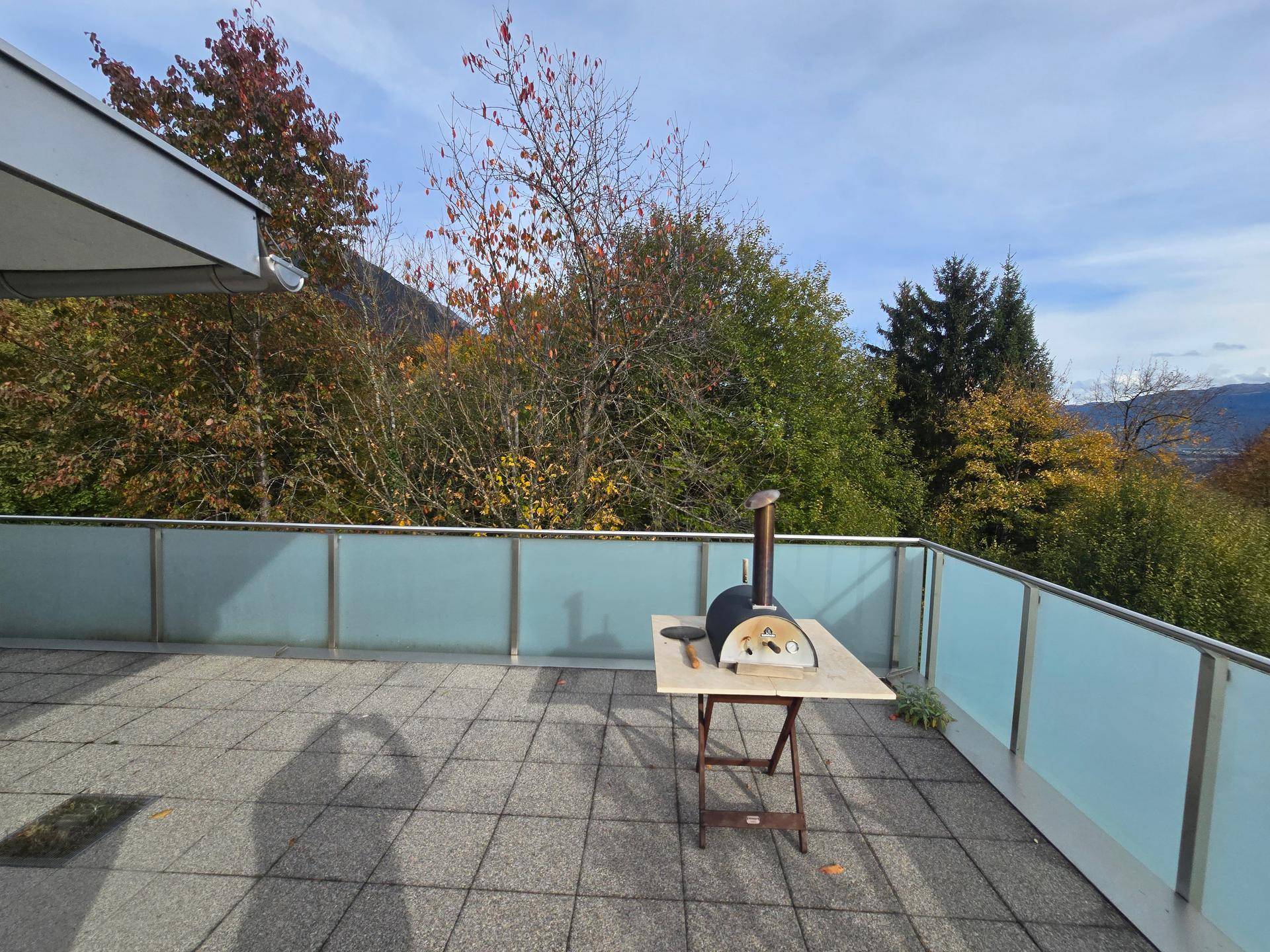 Großer Terrassenbereich mit Fliesenboden und einem Pizzaofen, umgeben von Bäumen.