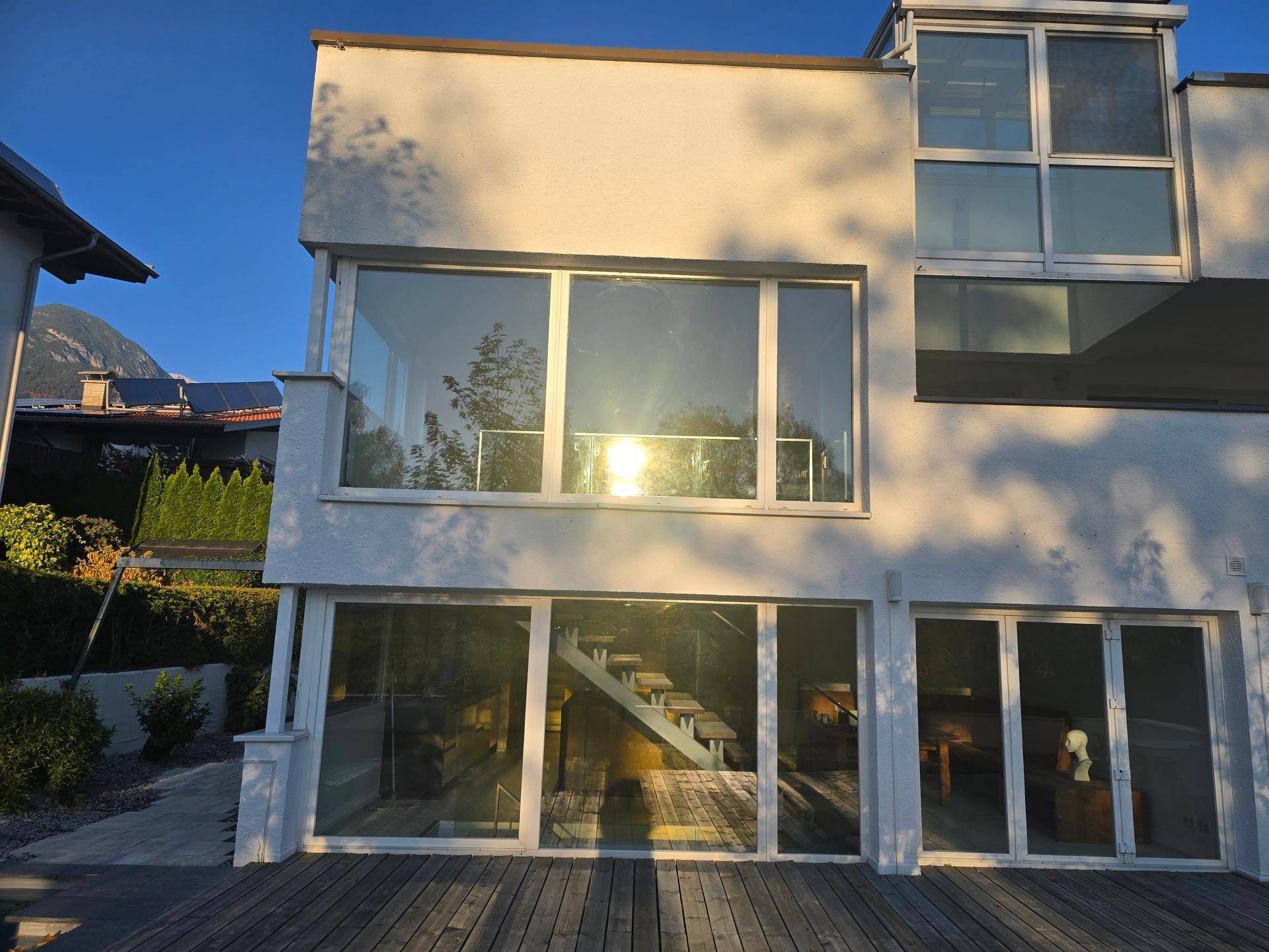 Moderne Hausfassade mit großen Glasflächen und sichtbarer Innentreppe im warmen Sonnenlicht.
