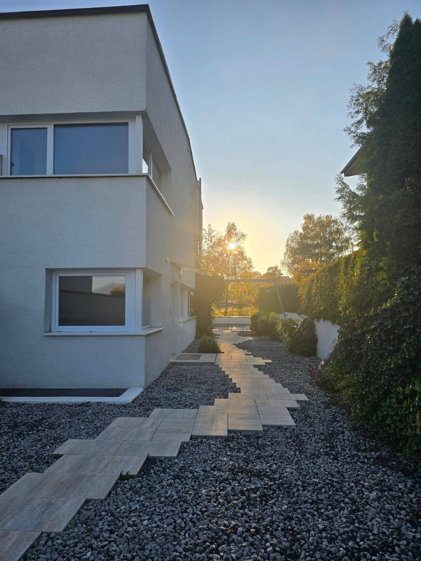 Seitenansicht des Hauses mit Gartenweg und Bäumen im warmen Licht der untergehenden Sonne.
