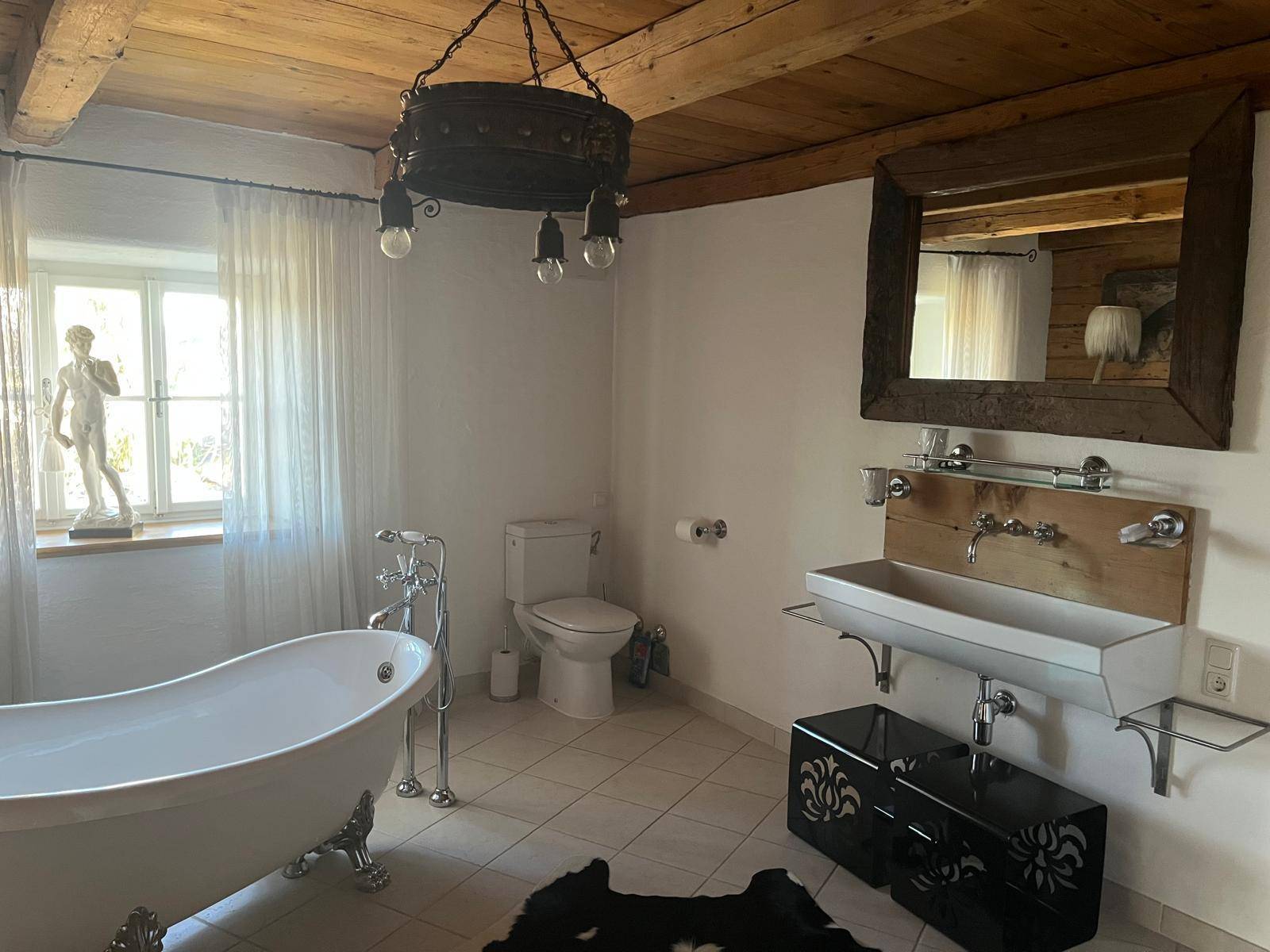 Helles Badezimmer mit freistehender Badewanne, Holzbalkendecke und rustikalem Spiegel über dem Waschbecken.