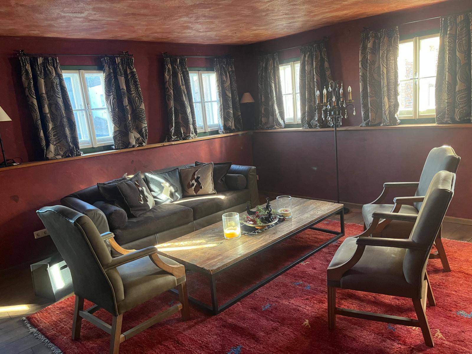 Gemütlicher Wohnbereich mit rotem Teppich, Holztisch und viel natürlichem Licht durch die Fenster.