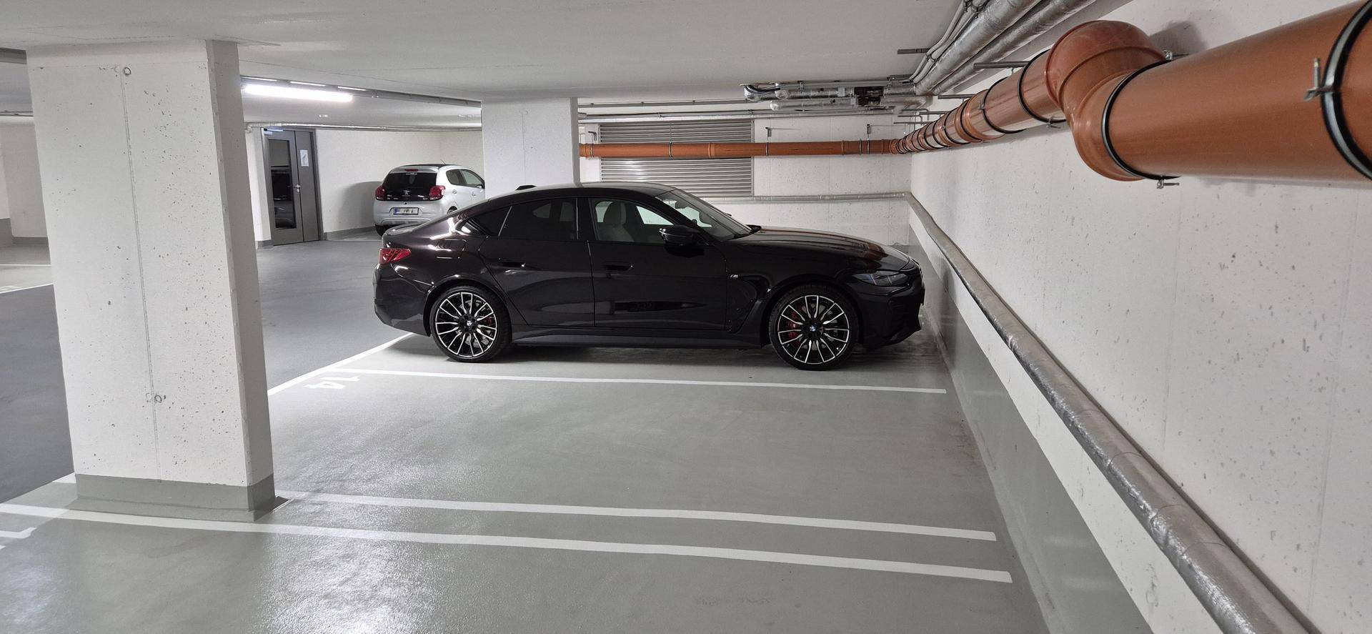 Schwarzes Auto auf einem Stellplatz in der Tiefgarage mit sichtbaren Rohrleitungen an der Wand.