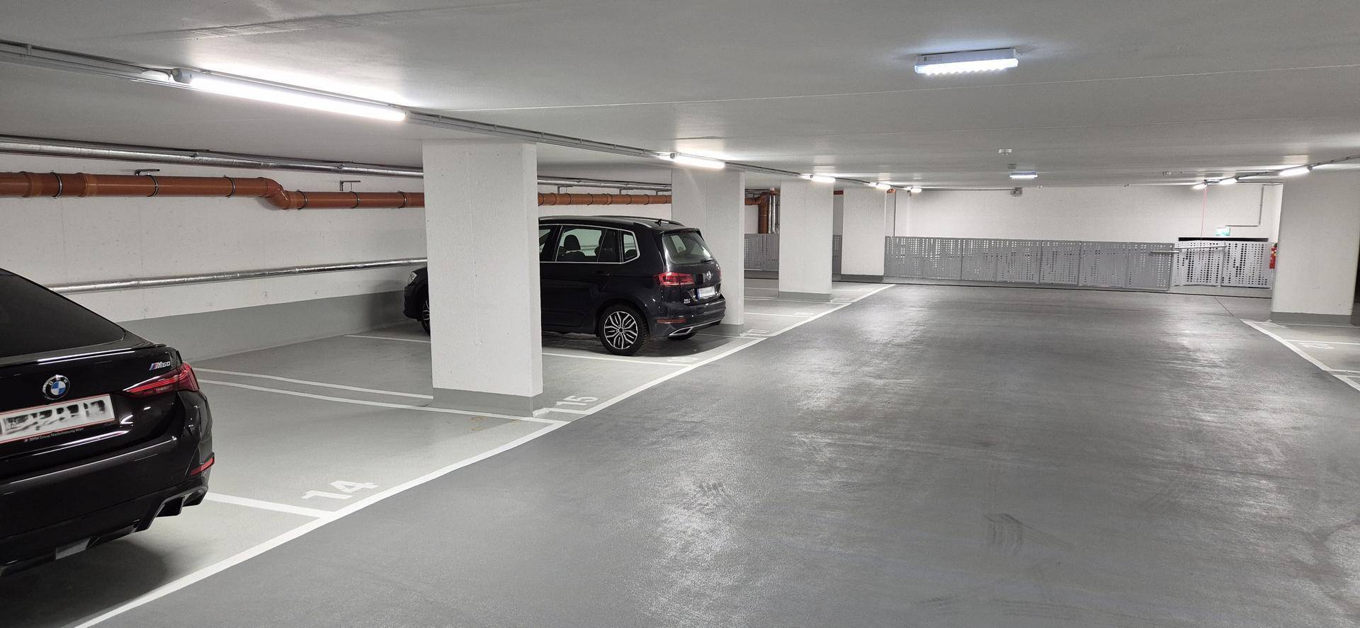 Tiefgarage mit geparkten Autos, sichtbaren Rohrleitungen an der Decke und heller Beleuchtung.