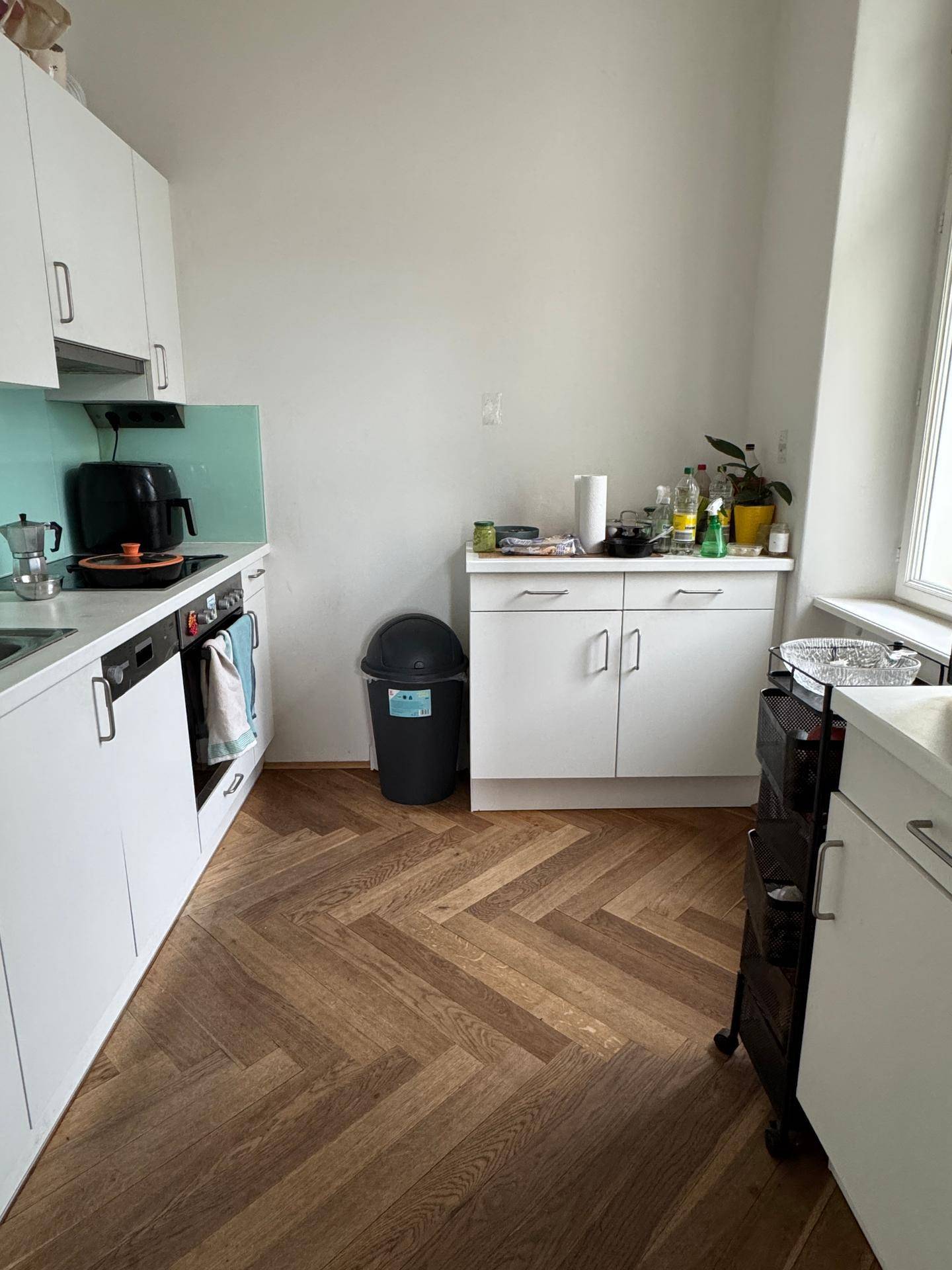 Funktionale Küche mit weißen Schränken, Fischgrätparkett und einem Fenster für Tageslicht.