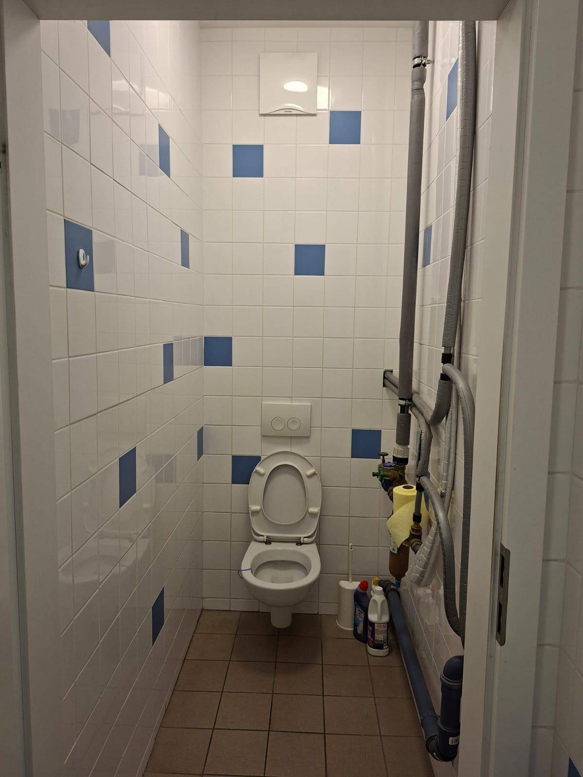 Kleine Toilette mit weißen und blauen Fliesen, Wand-WC und sichtbaren Installationsrohren an der Wand.