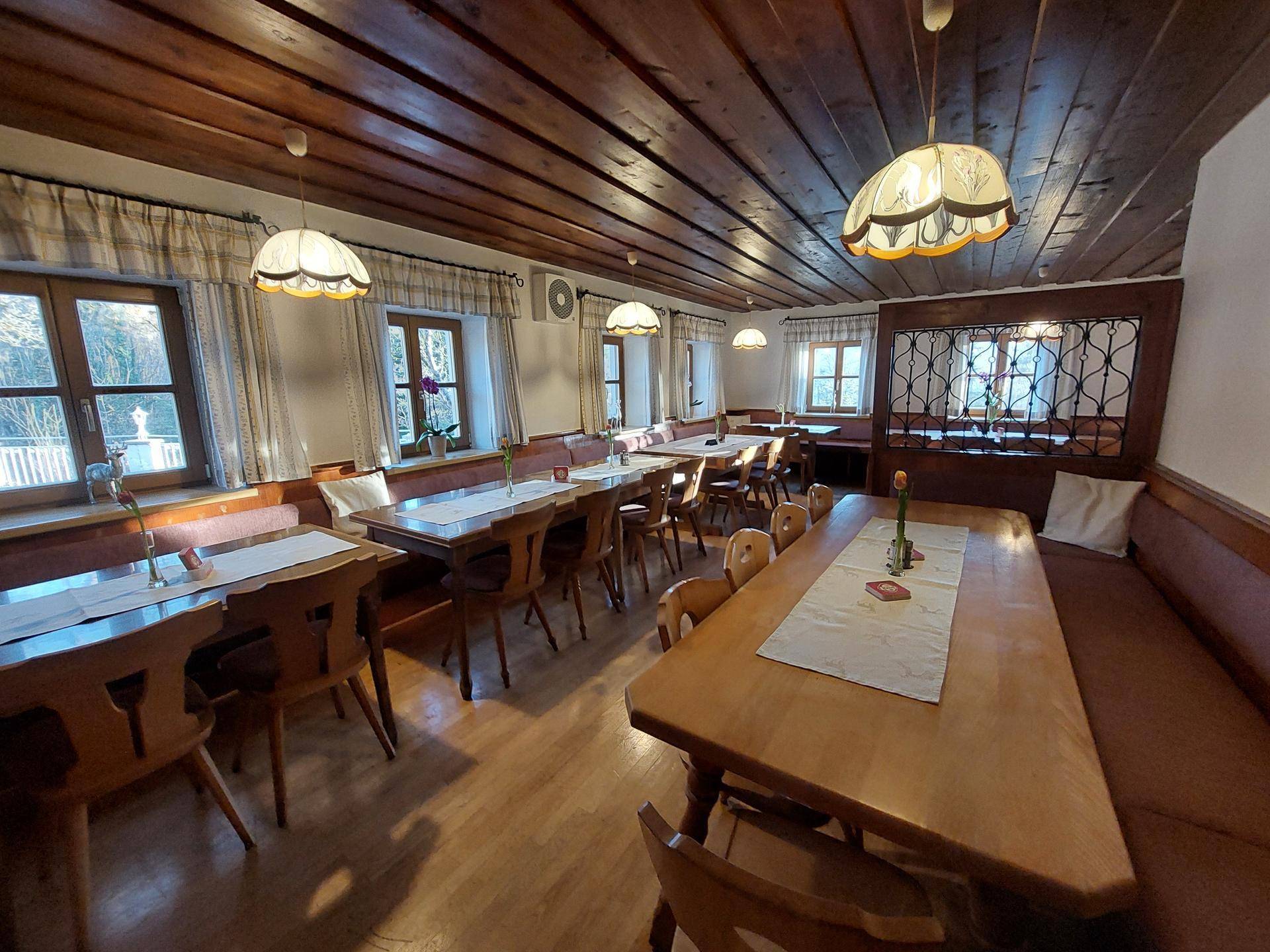 Rustikaler Speisesaal mit dunkler Holzbalkendecke, Holztischen und Bänken, beleuchtet durch mehrere Fenster.