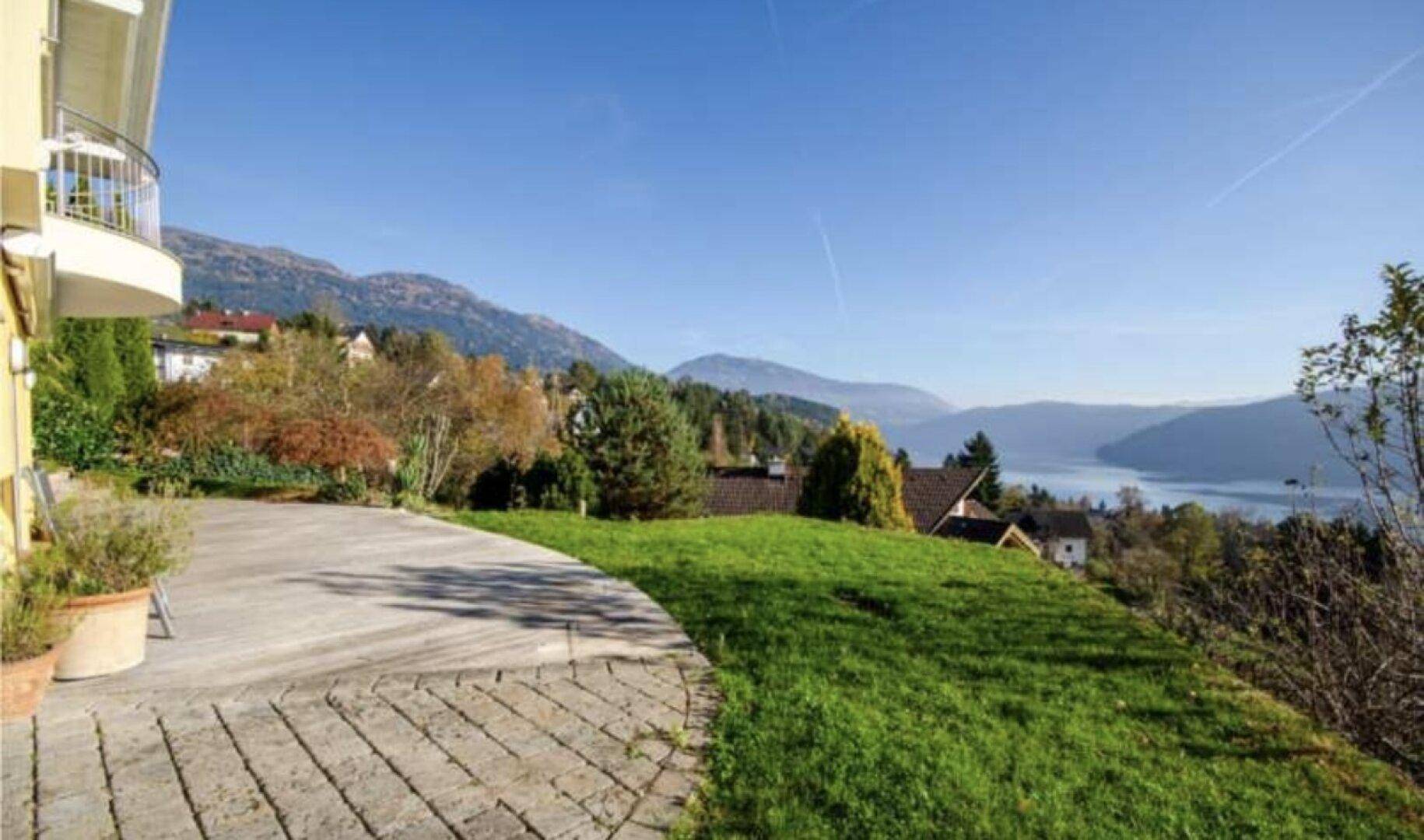 Gepflasterte Terrasse neben dem Haus mit Rasenfläche und weitem Blick auf den See und die Berge.