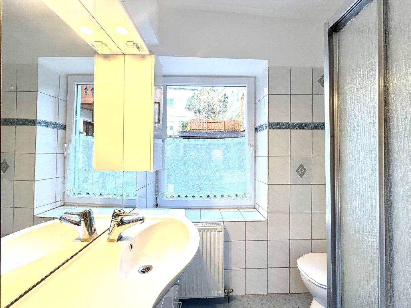 Helles Badezimmer mit Fenster, Waschbecken, Spiegel und Duschkabine.