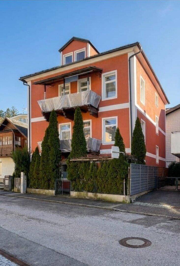 Traditionelles Gebäude mit orangefarbener Fassade, Balkon und hohen Bäumen an der Straße.