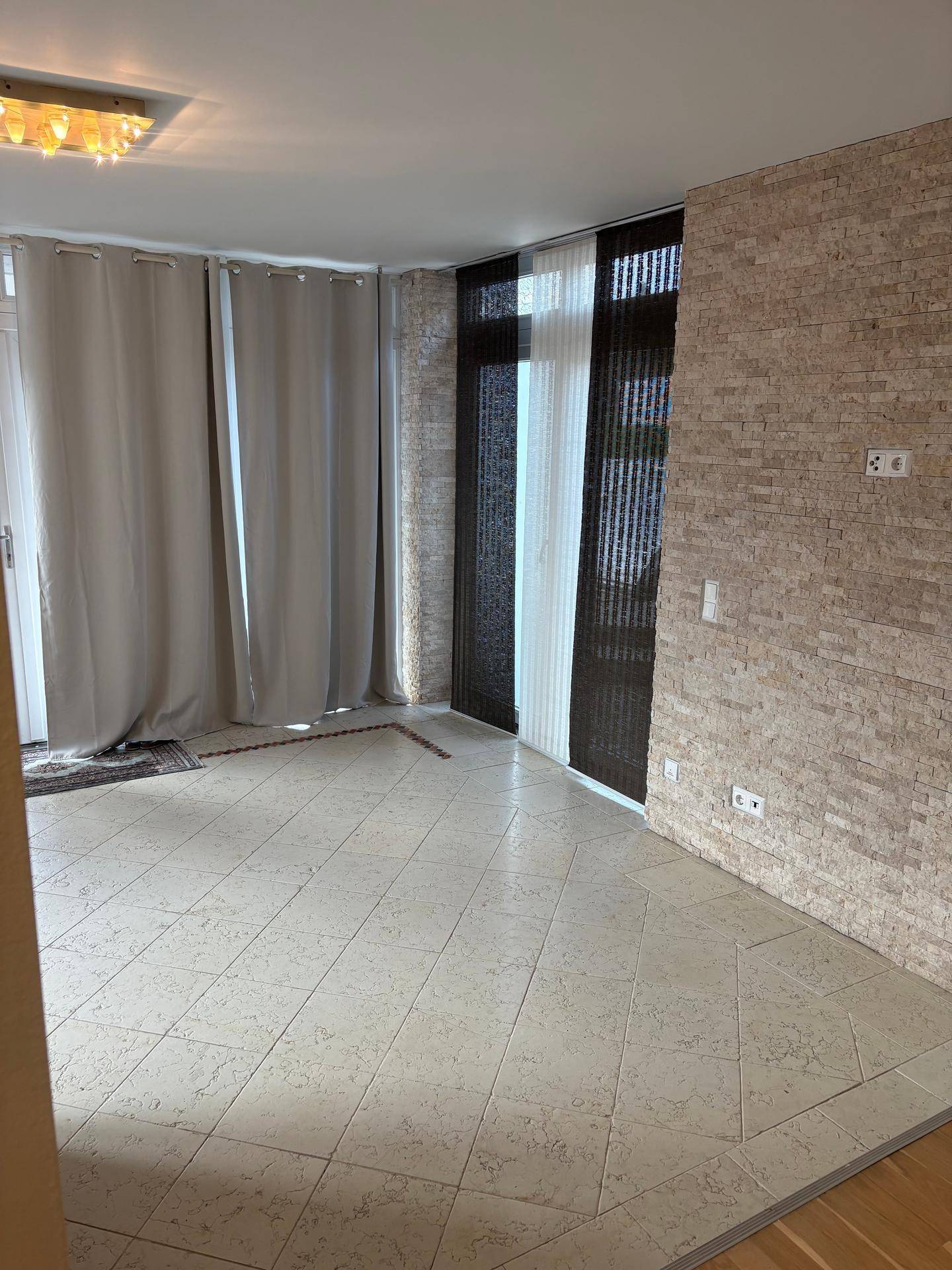 Innenraum mit dekorativer Steinwand, Fliesenboden und Vorhängen an der Terrassentür.