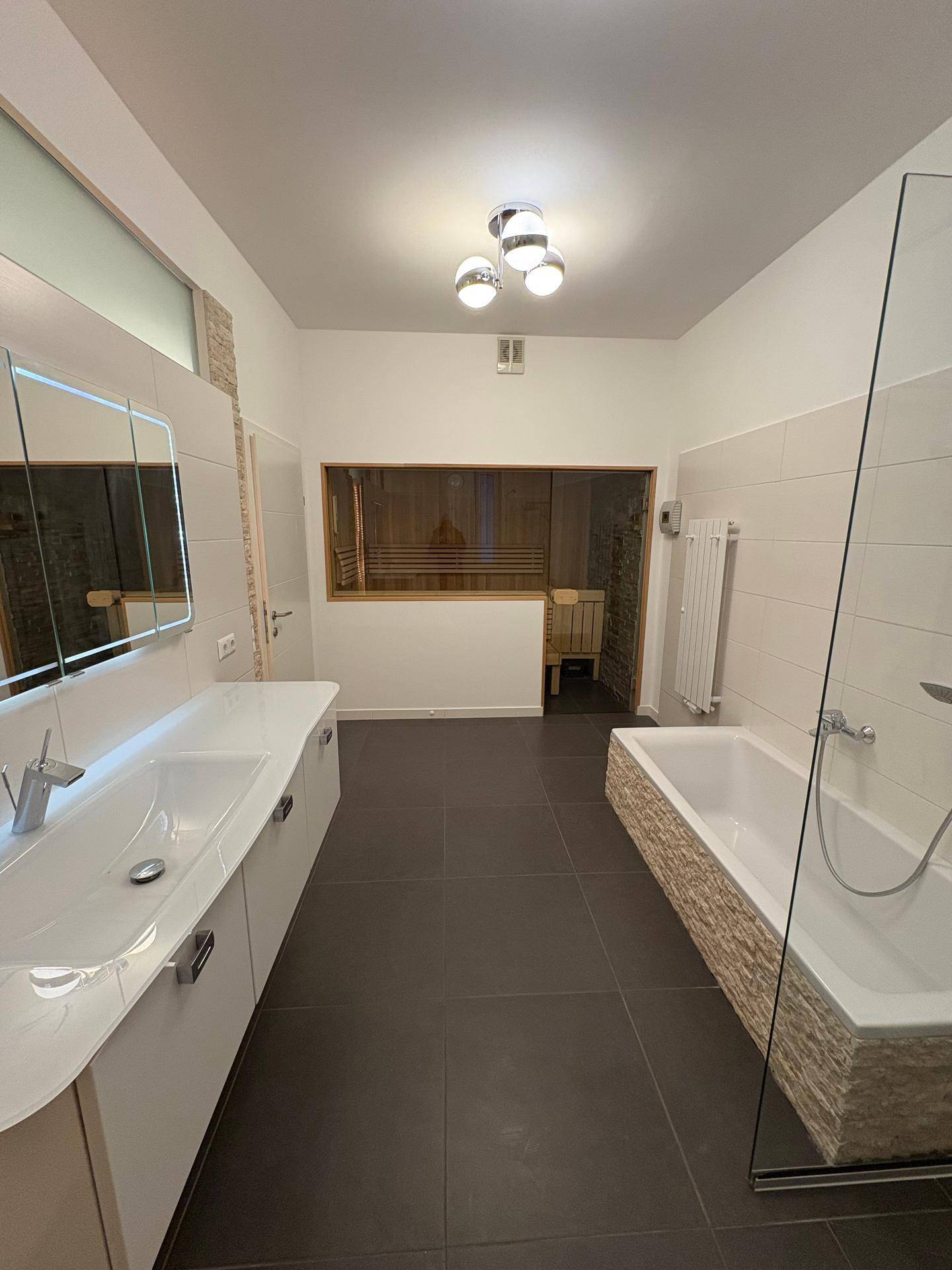 Modernes Badezimmer mit Doppelwaschbecken, Badewanne und integrierter Sauna im Hintergrund.