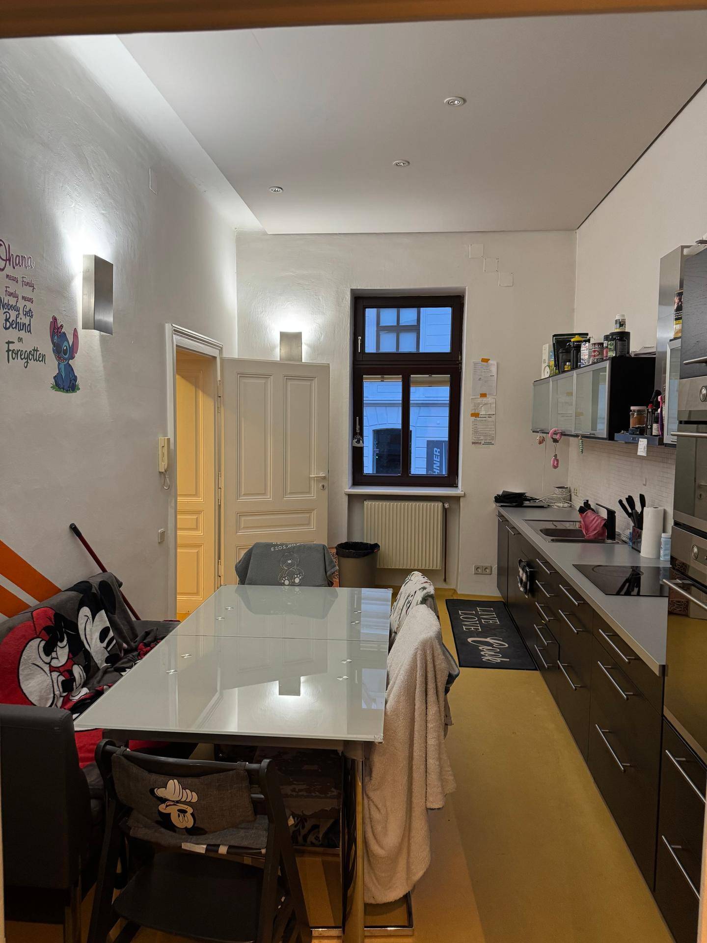 Lange Küche mit Esstisch, dunklen Schränken und einem Fenster.